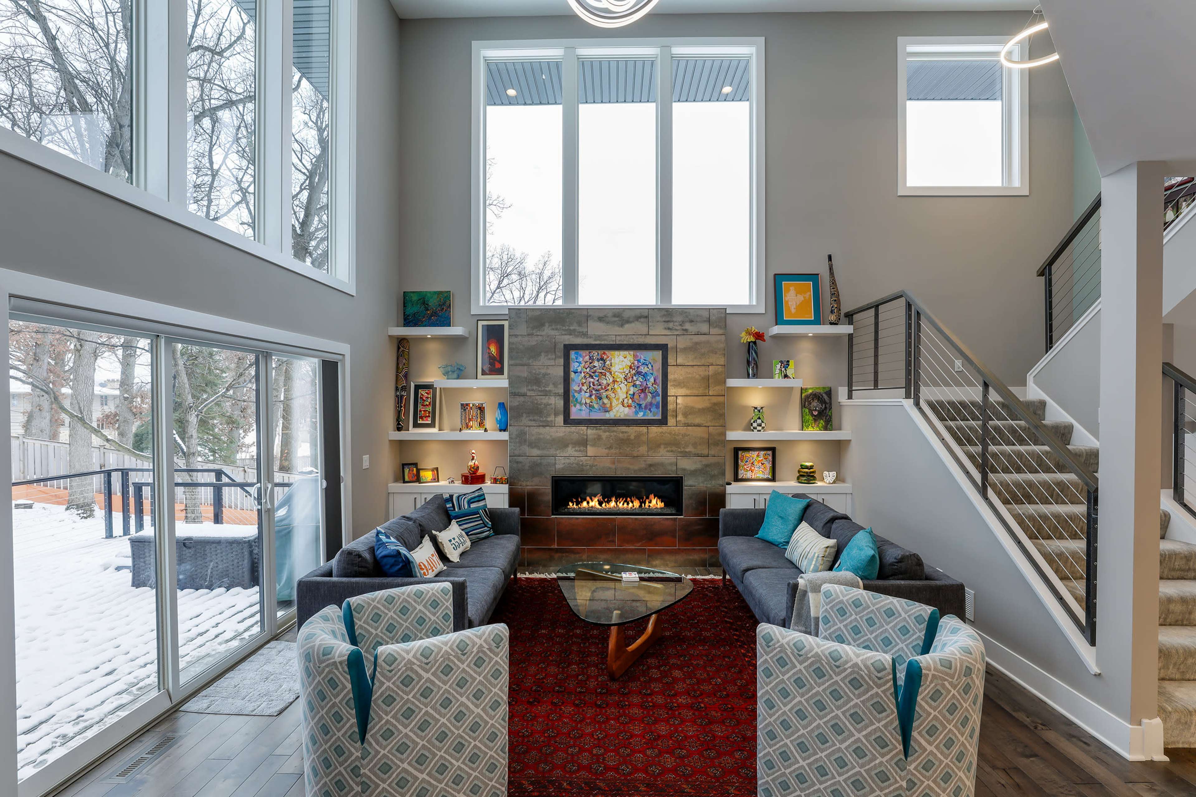 A modern living room with large windows, a central fireplace, an area rug, and a staircase leading to an upper level.
