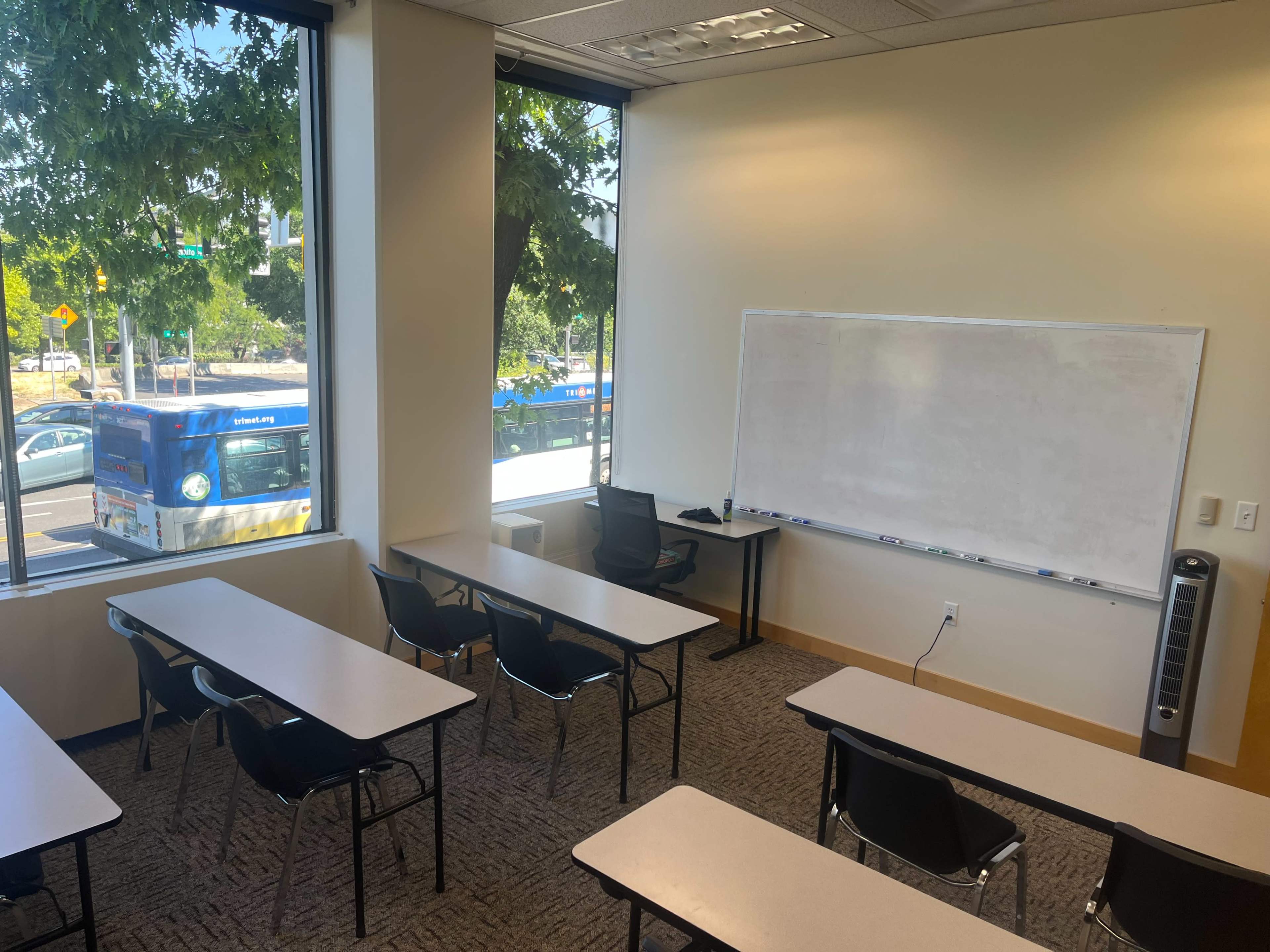 The image shows a classroom with rows of tables and chairs, a whiteboard on the wall, and large windows revealing a view of a street with a bus passing by.