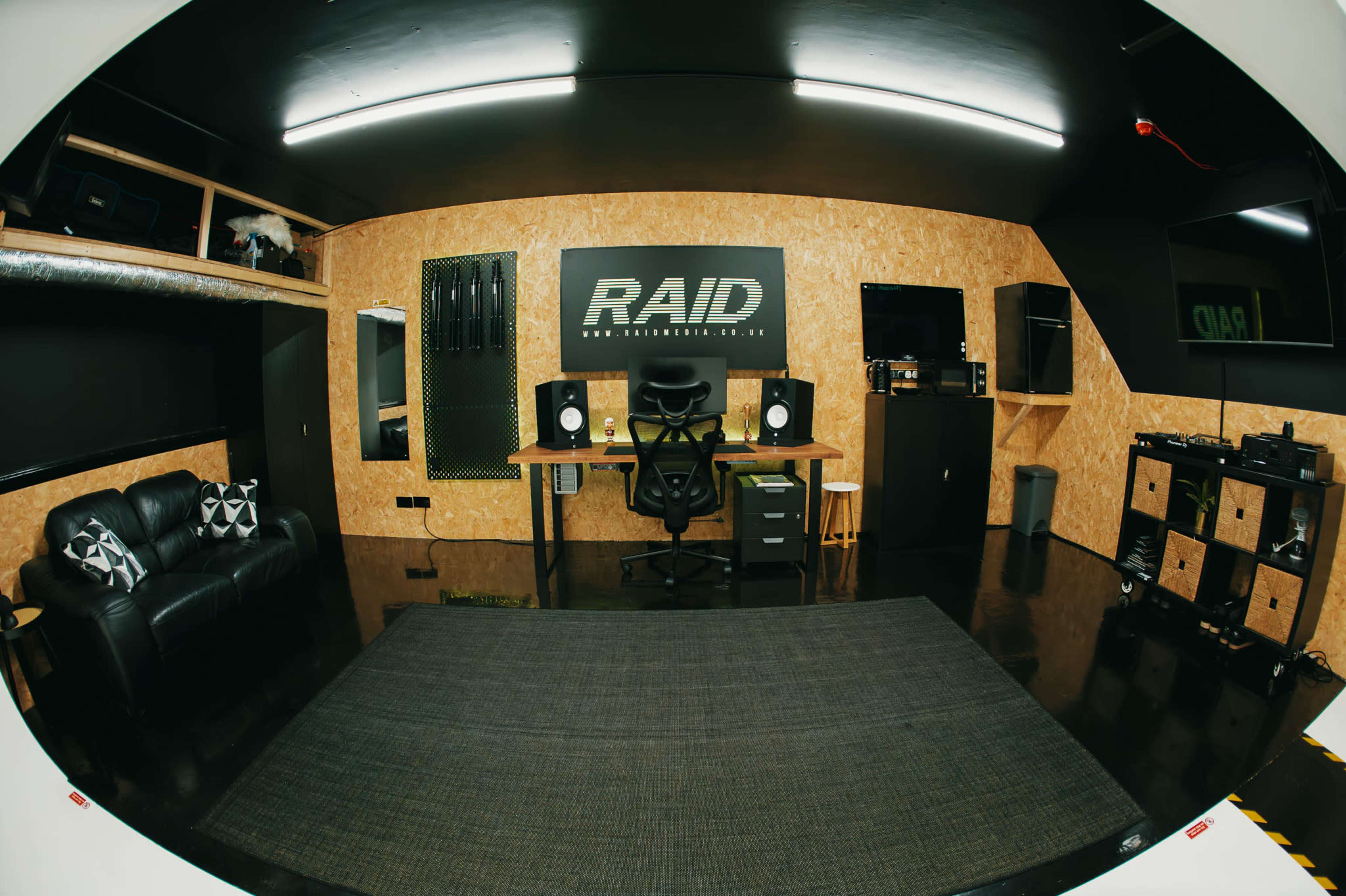 The image shows a modern studio setup featuring a desk with an office chair, a sofa, and several sound equipment units against a wooden wall with a RAID logo.