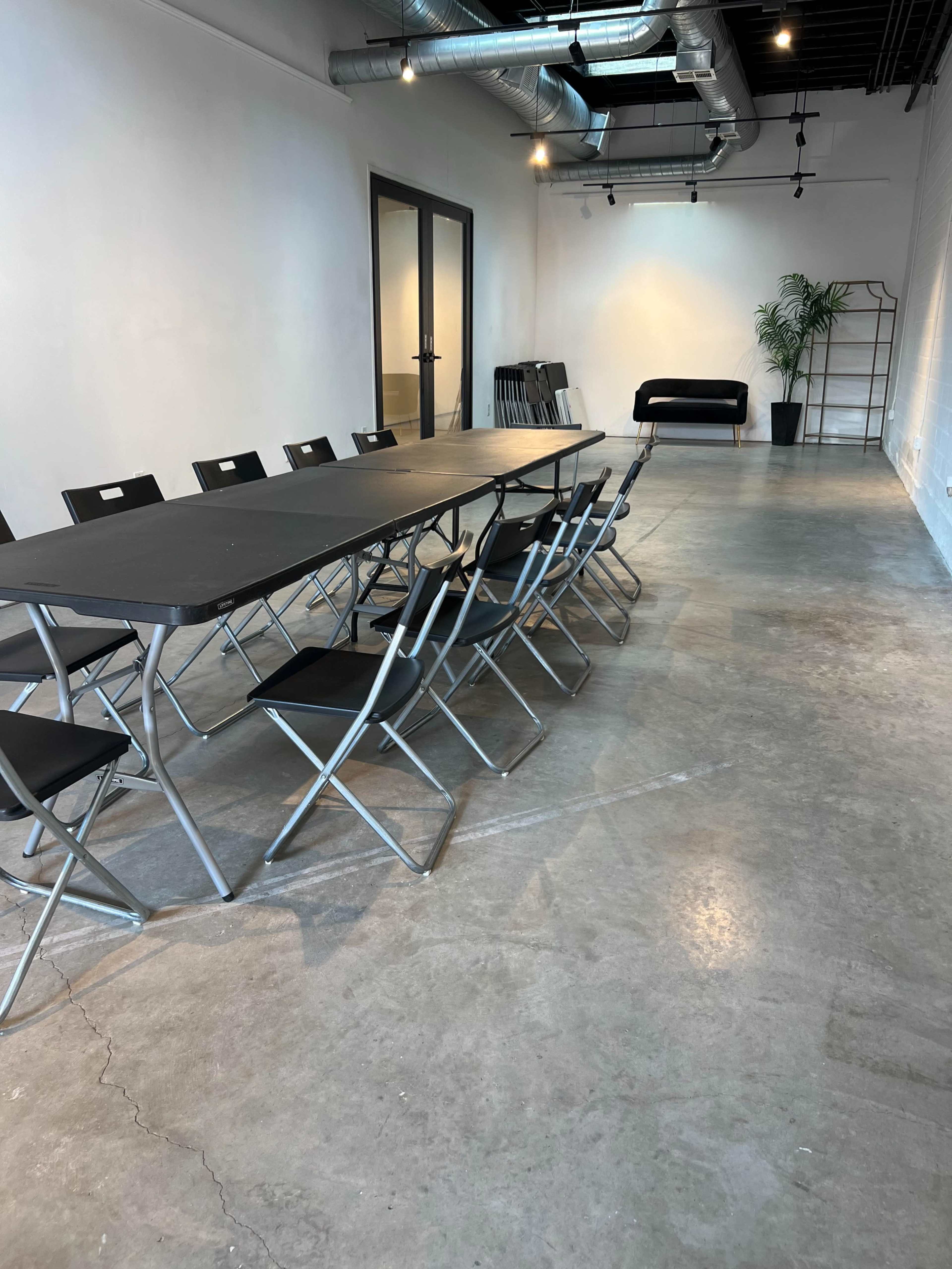 The image shows a minimalist conference room with a long table, several folding chairs, and a black sofa against the wall.
