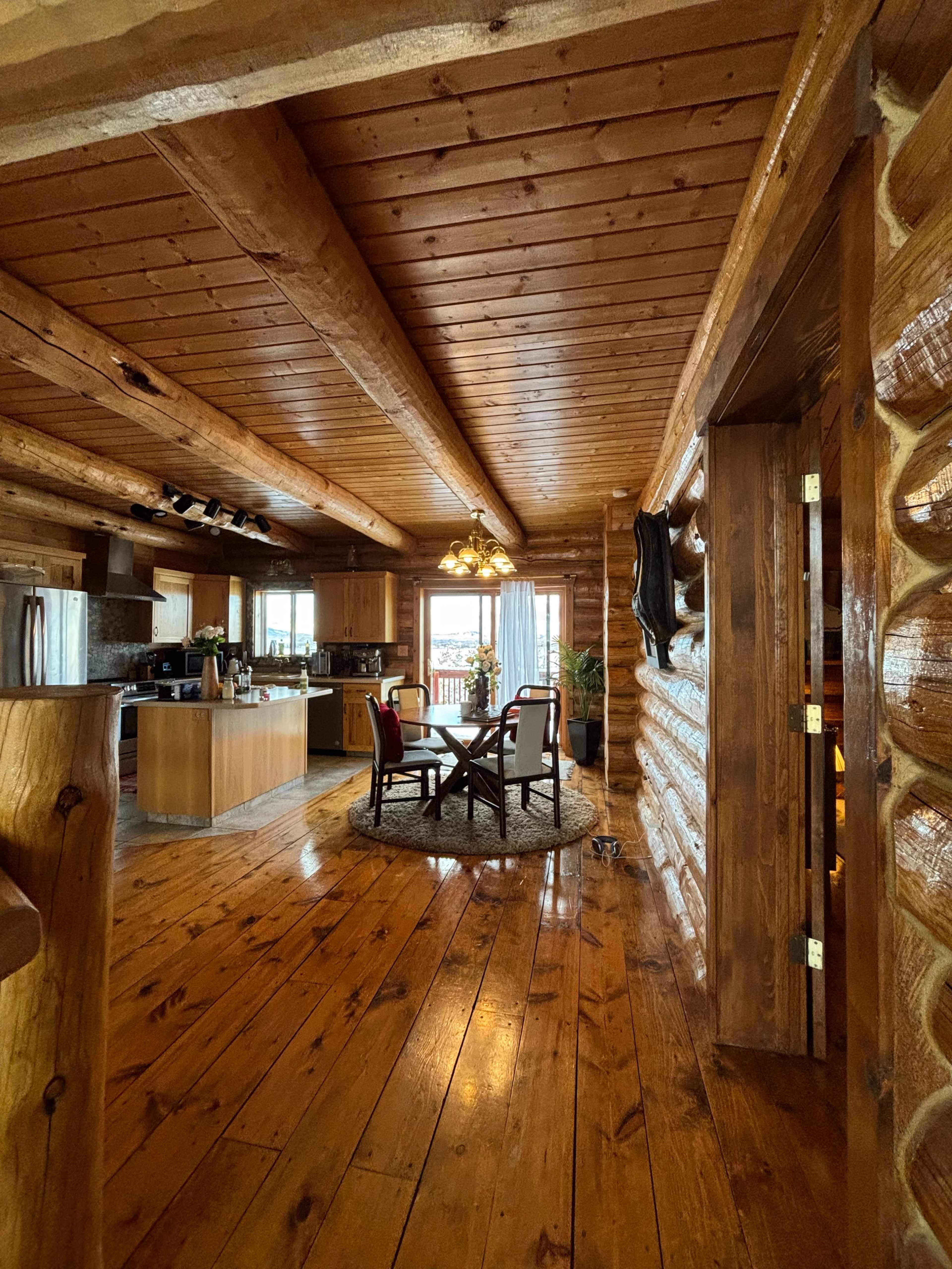 The image shows a wooden interior of a log cabin featuring an open kitchen and dining area with a circular table and chairs.
