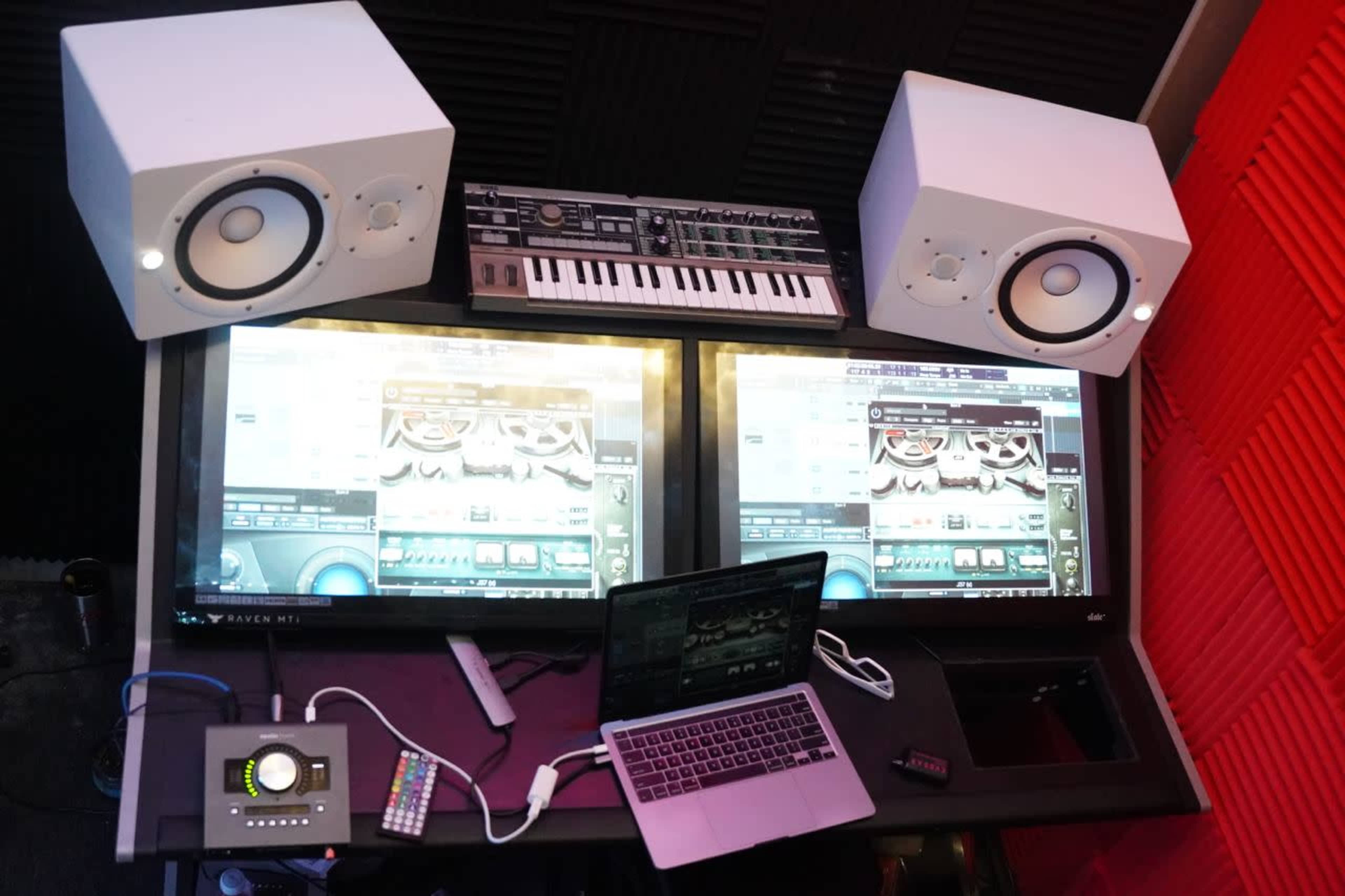 A music production setup featuring two large monitors, a keyboard, and studio monitors on a desk with a laptop and audio control interface.