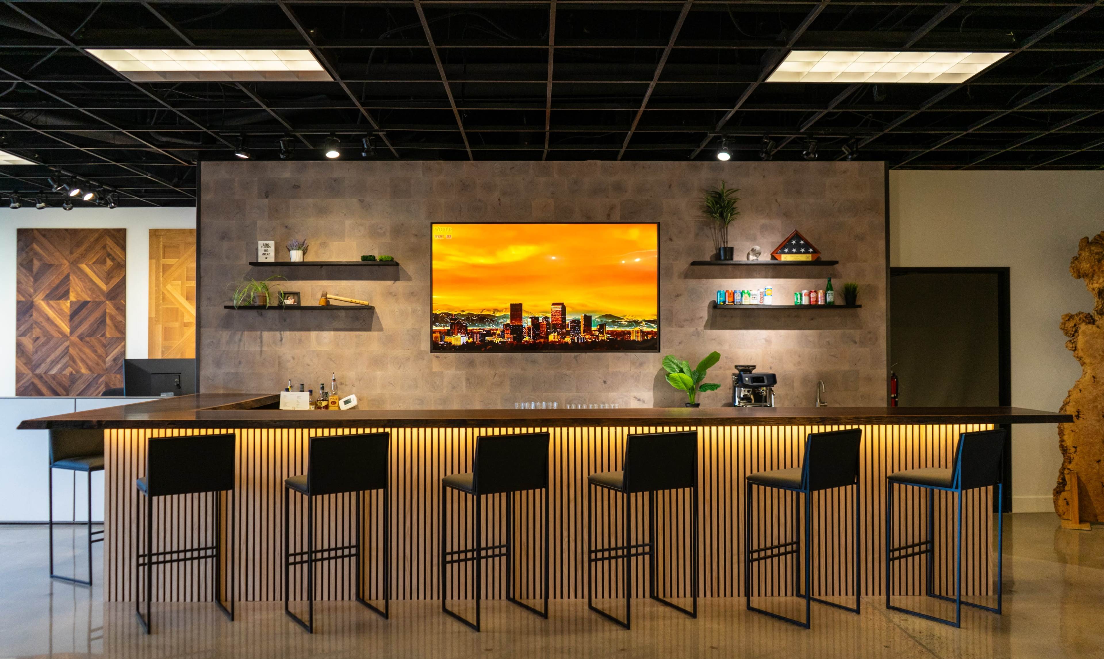 The image shows a modern bar area with a sleek counter, backlit with warm lighting, and a large television mounted on a textured wall.