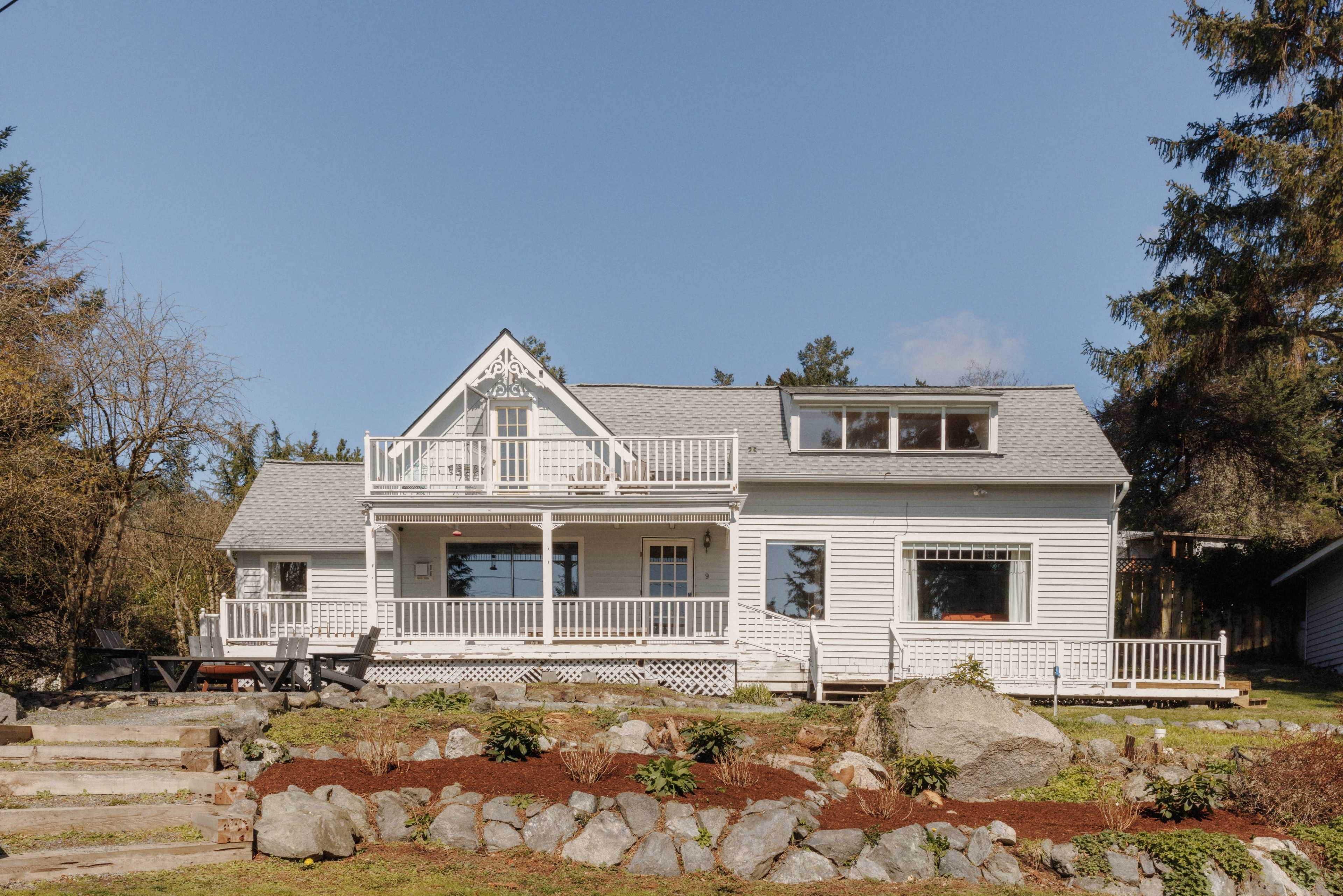 A large, white, two-story house with a front porch and balcony is surrounded by a landscaped yard featuring rocks and greenery.
