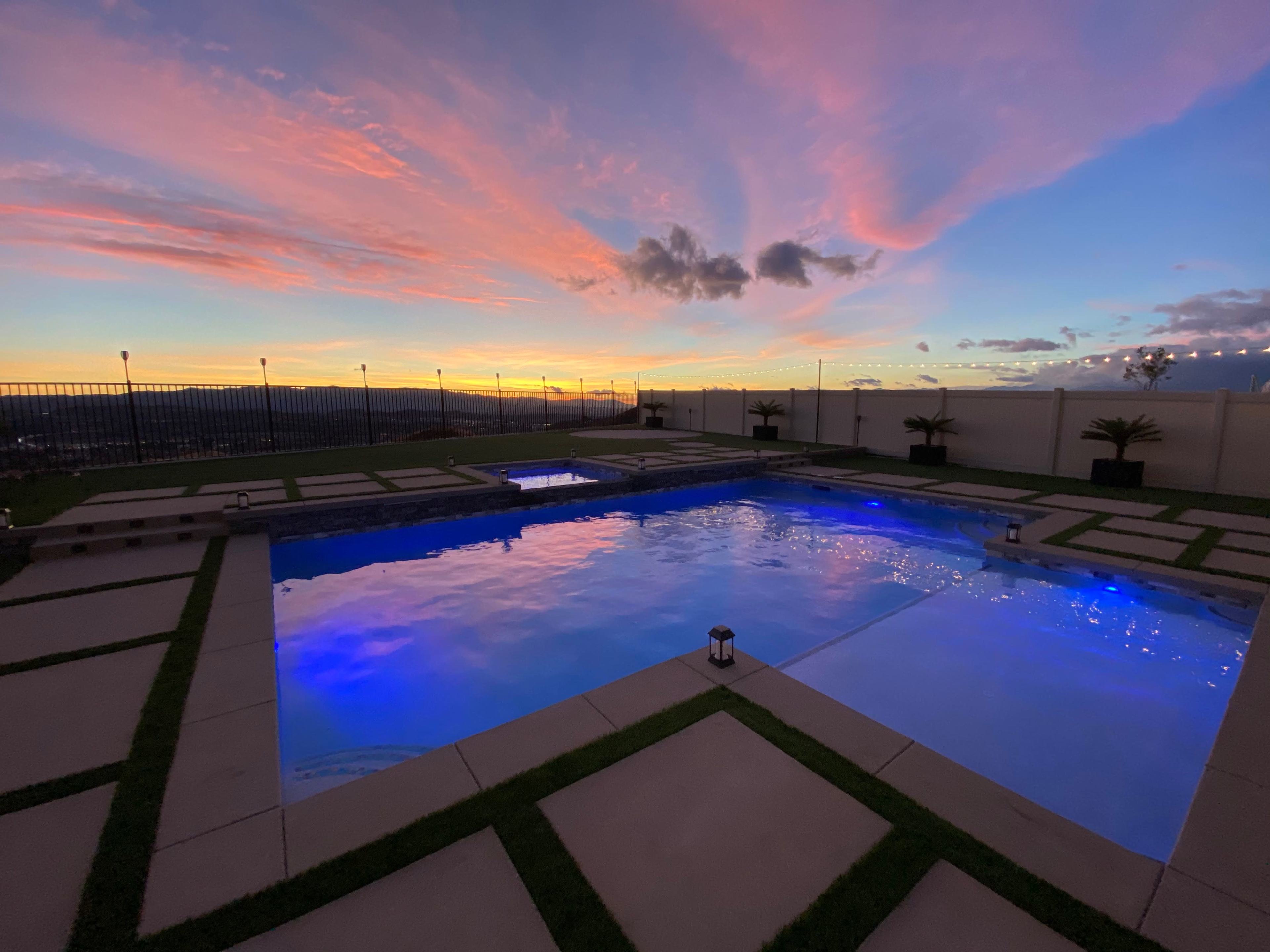 A swimming pool illuminated by blue lights sits surrounded by paved landscaping under a colorful sunset sky.