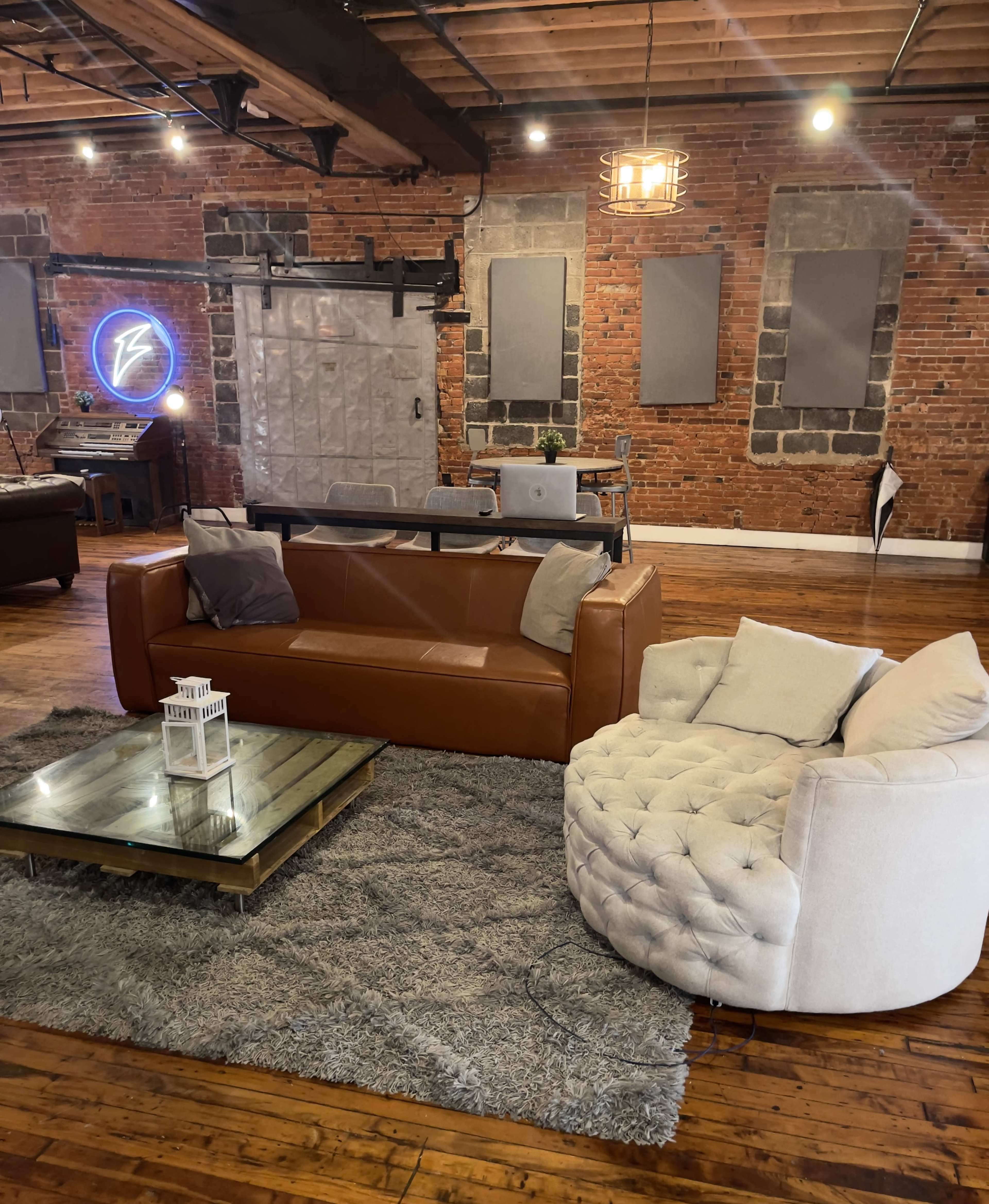 The image shows a modern living space with a brown leather sofa, a round white tufted chair, a glass coffee table on a gray rug, and exposed brick walls.