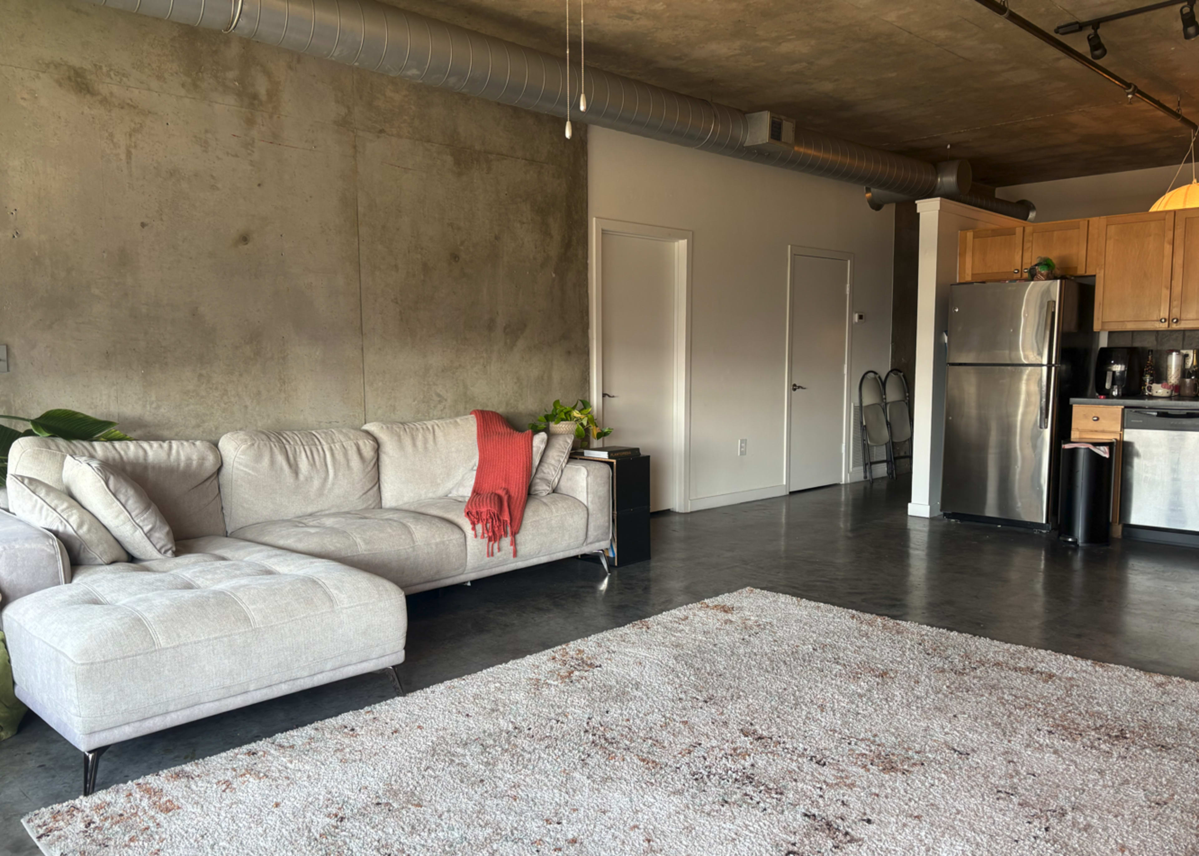 The image shows a modern living space featuring a light-colored sectional sofa, a large area rug, and an adjacent kitchen with stainless steel appliances.