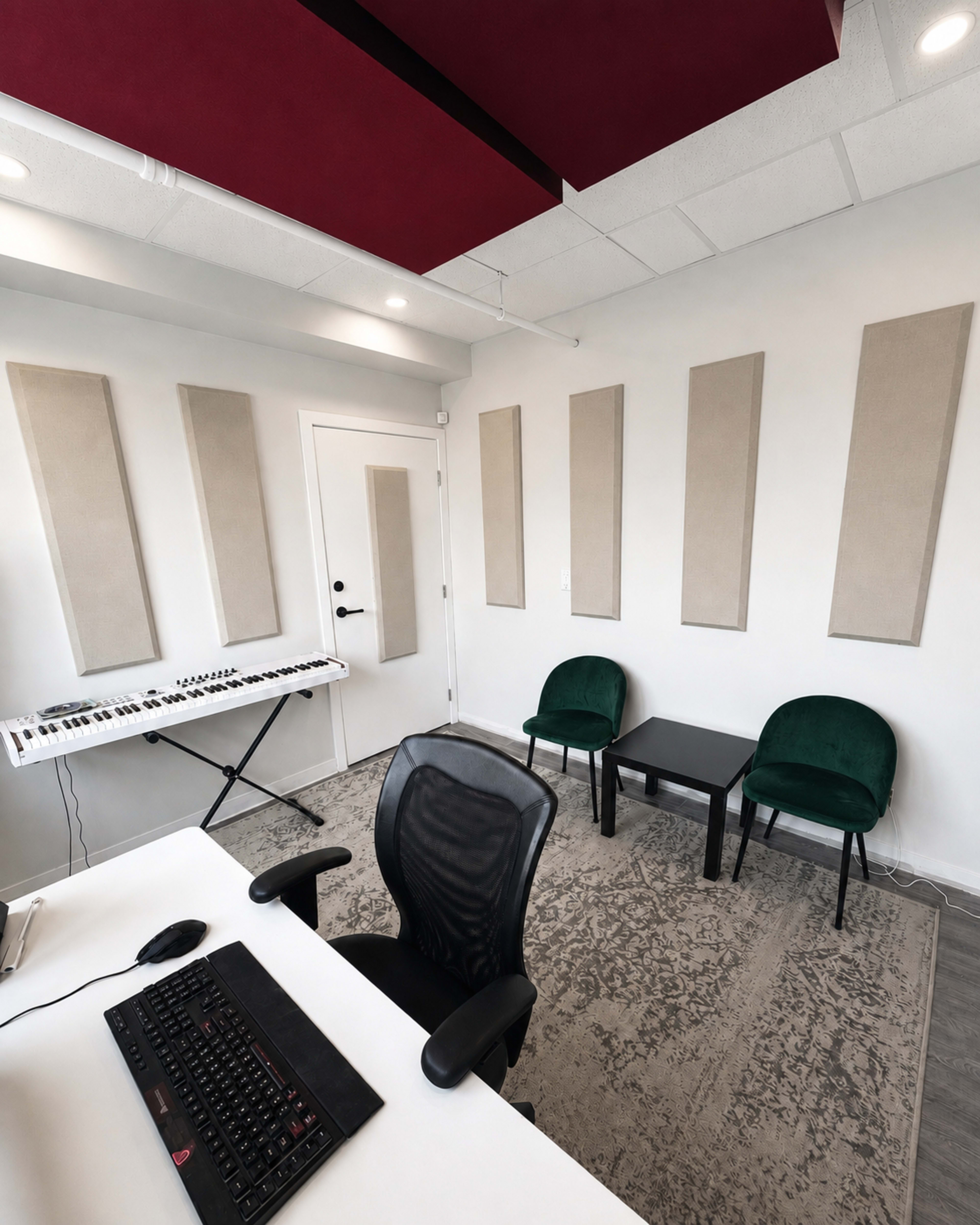 The image shows a music studio with a keyboard on a white stand, two green chairs, a small black table, and sound-absorbing panels on the walls.