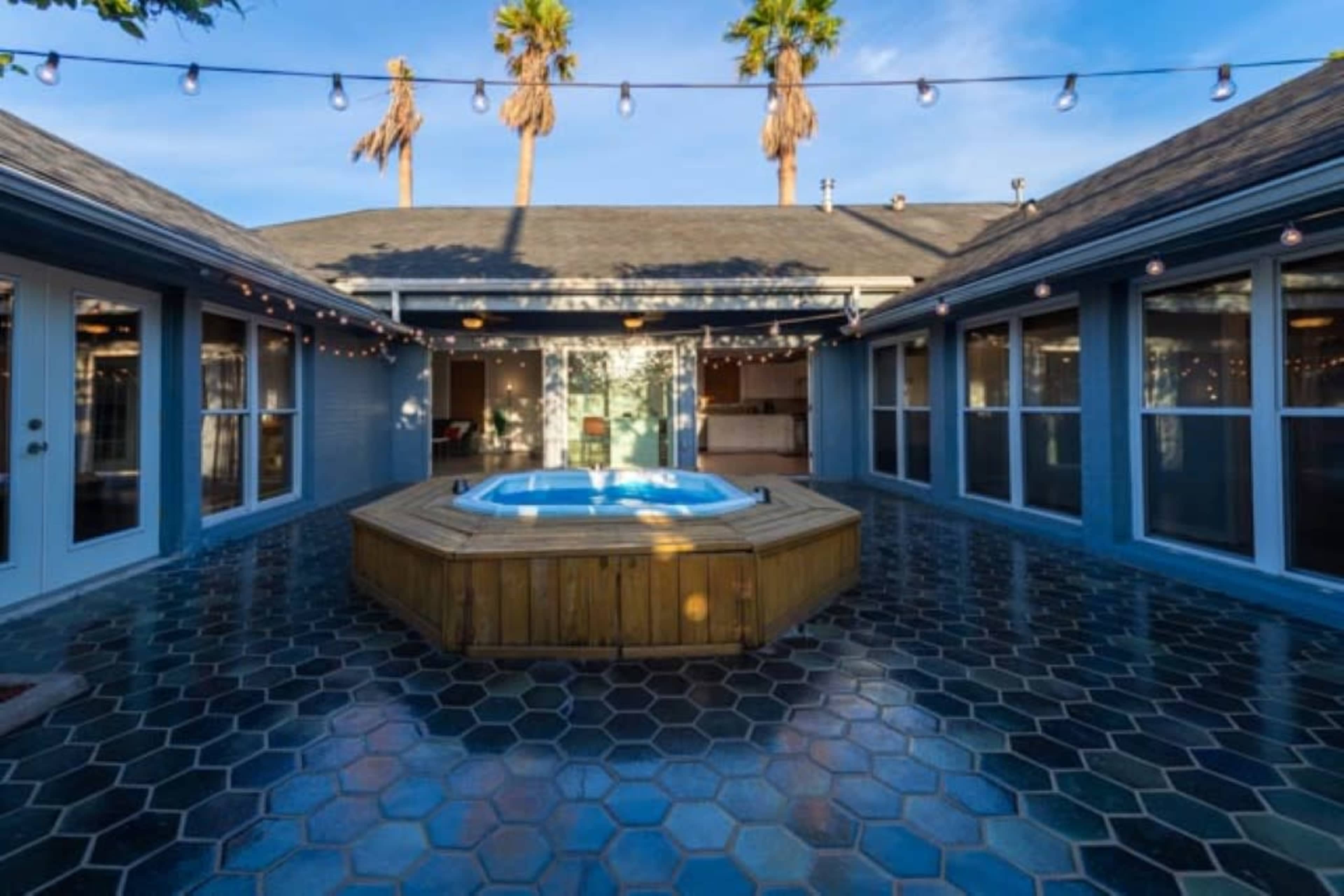 A wooden hot tub is situated in a courtyard surrounded by a tiled patio and enclosed by sliding glass doors, with string lights overhead and palm trees in the background.