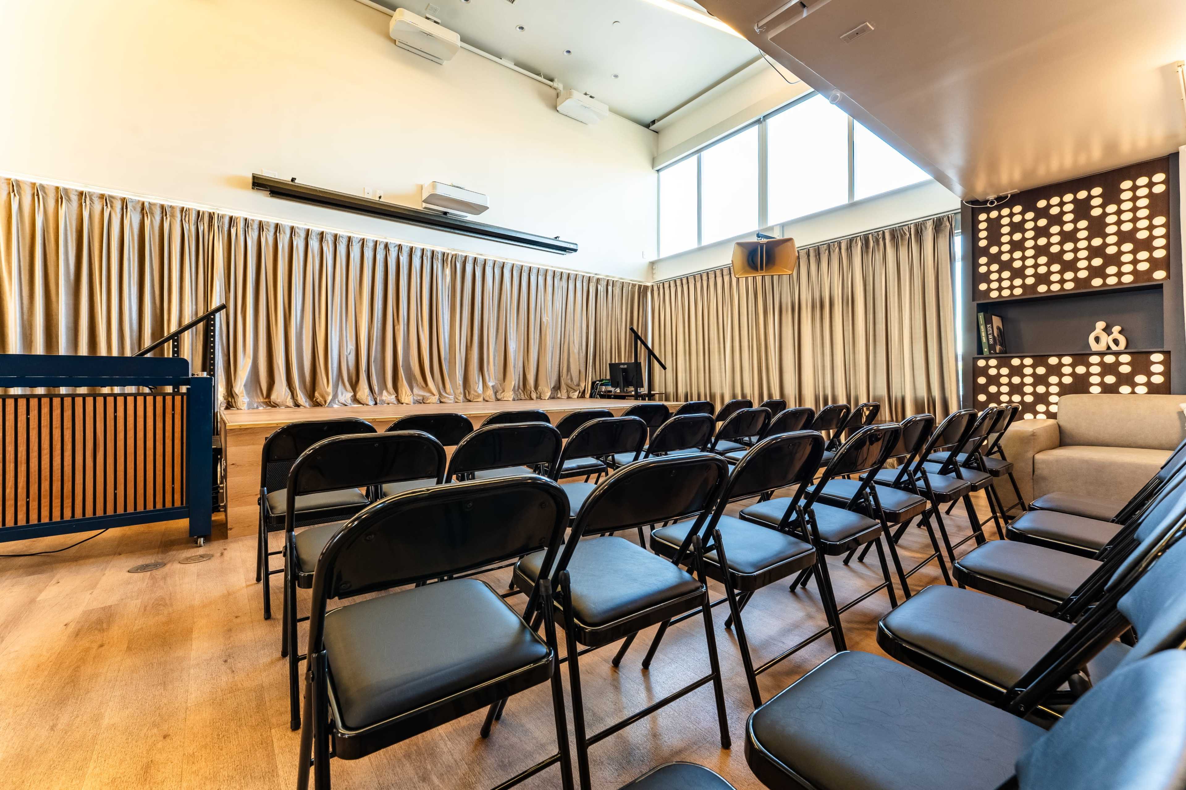The image shows a room with rows of black folding chairs facing a small stage, flanked by heavy curtains and featuring a wall with decorative lights.