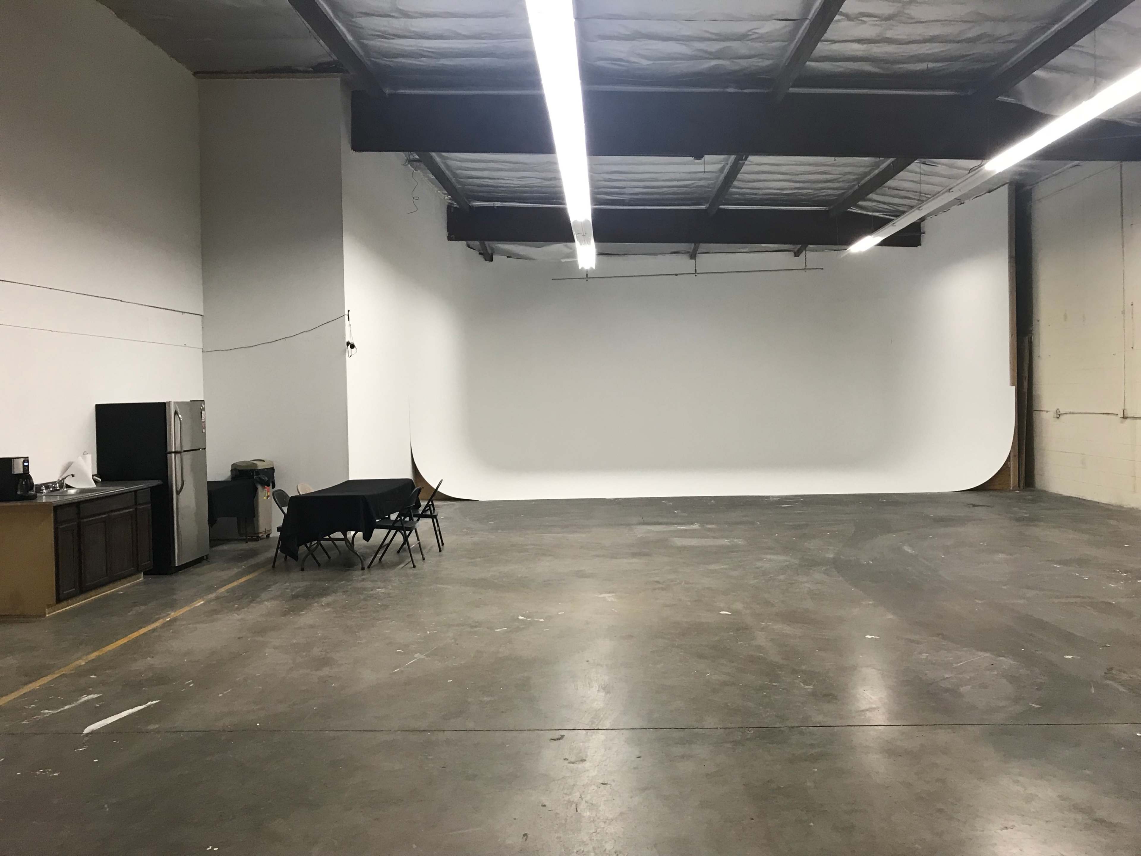 A spacious, empty industrial studio with a white backdrop, a kitchenette in the corner, and a few black folding chairs set up nearby.