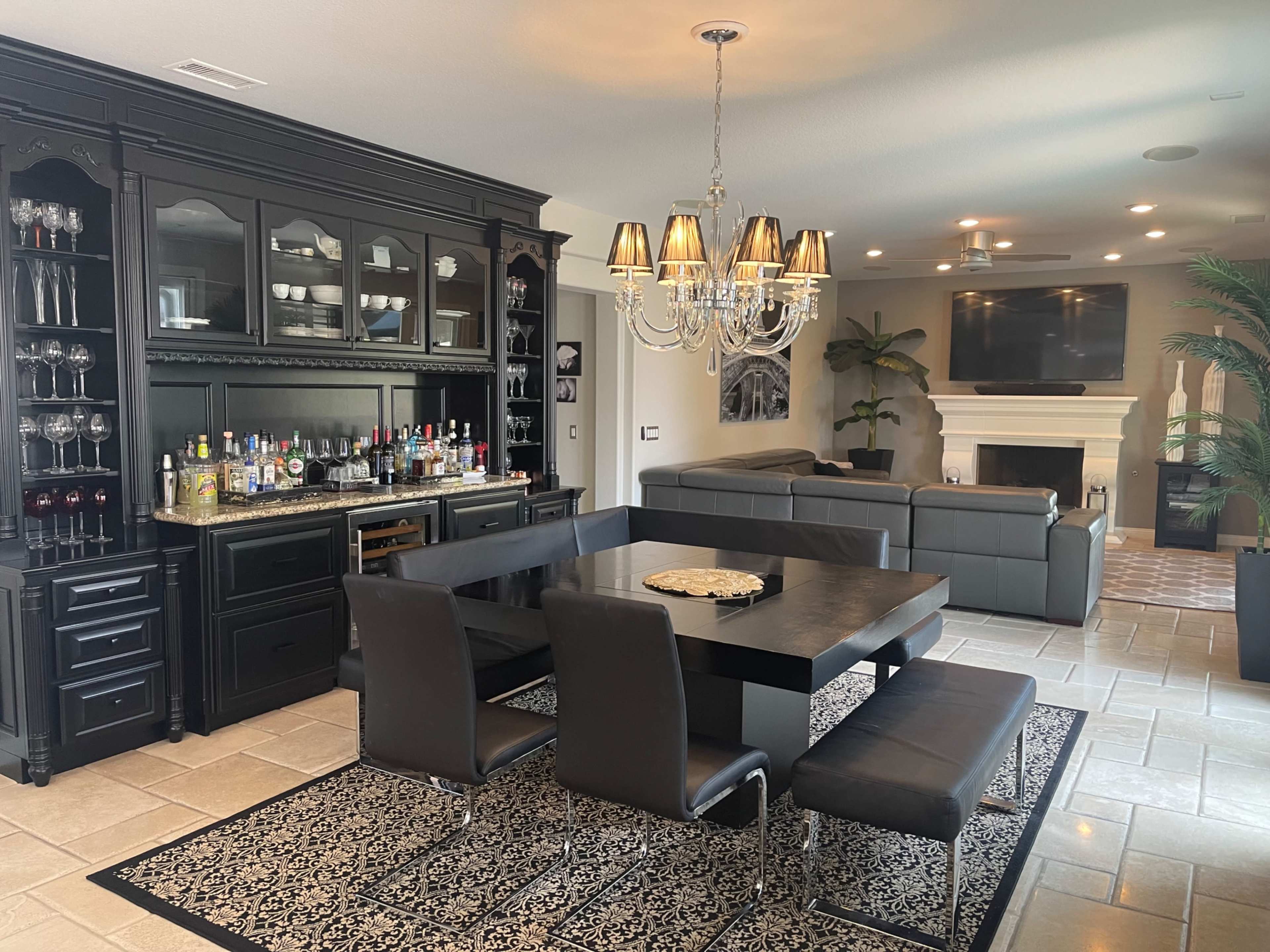 A dining area features a black dining table surrounded by chairs, with a bar cabinet filled with glassware and a chandelier overhead, adjacent to a living space with a sofa and a fireplace.