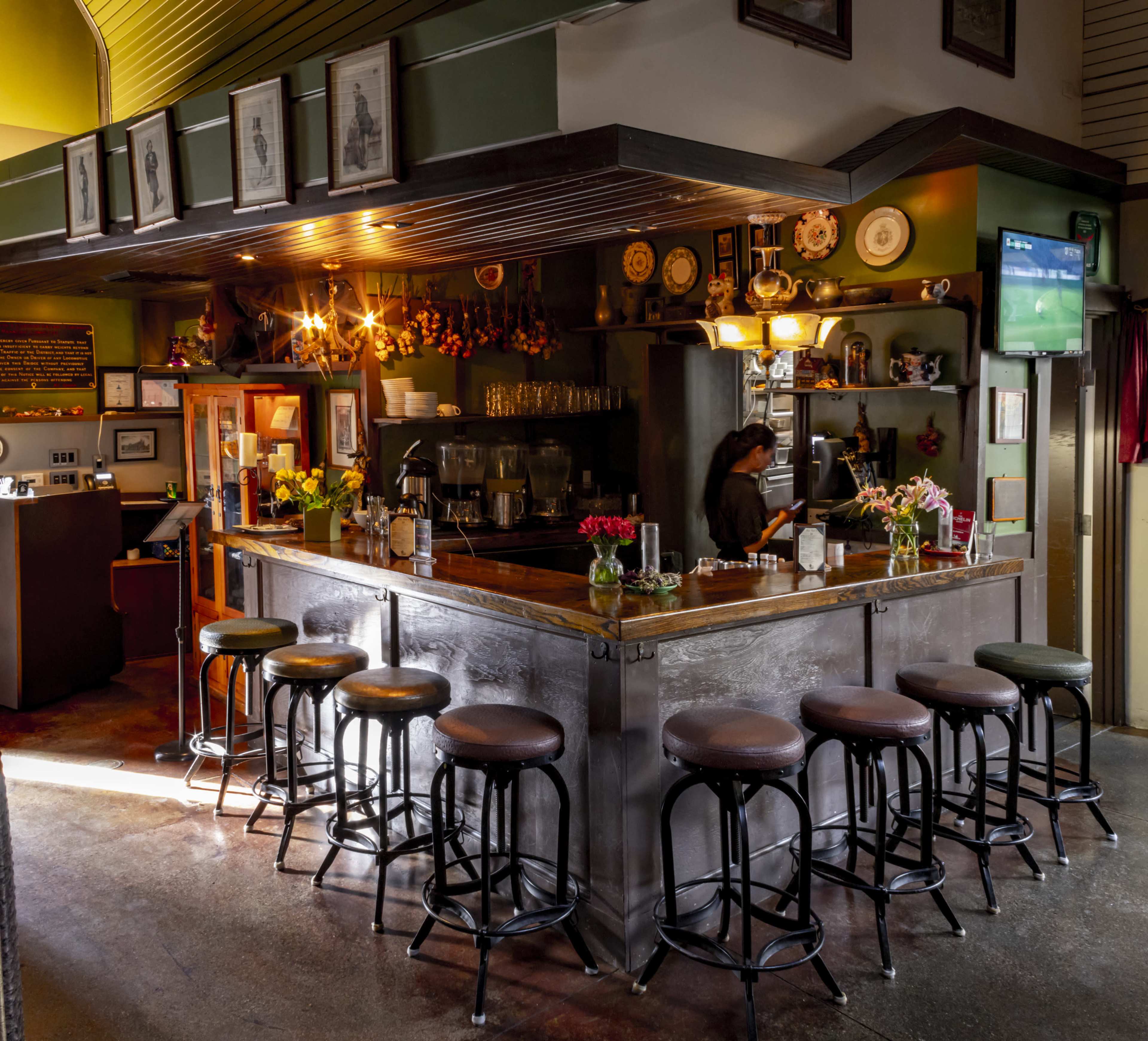 A cozy bar with a wooden counter, several stools, and a backdrop adorned with photographs and decorative plates.