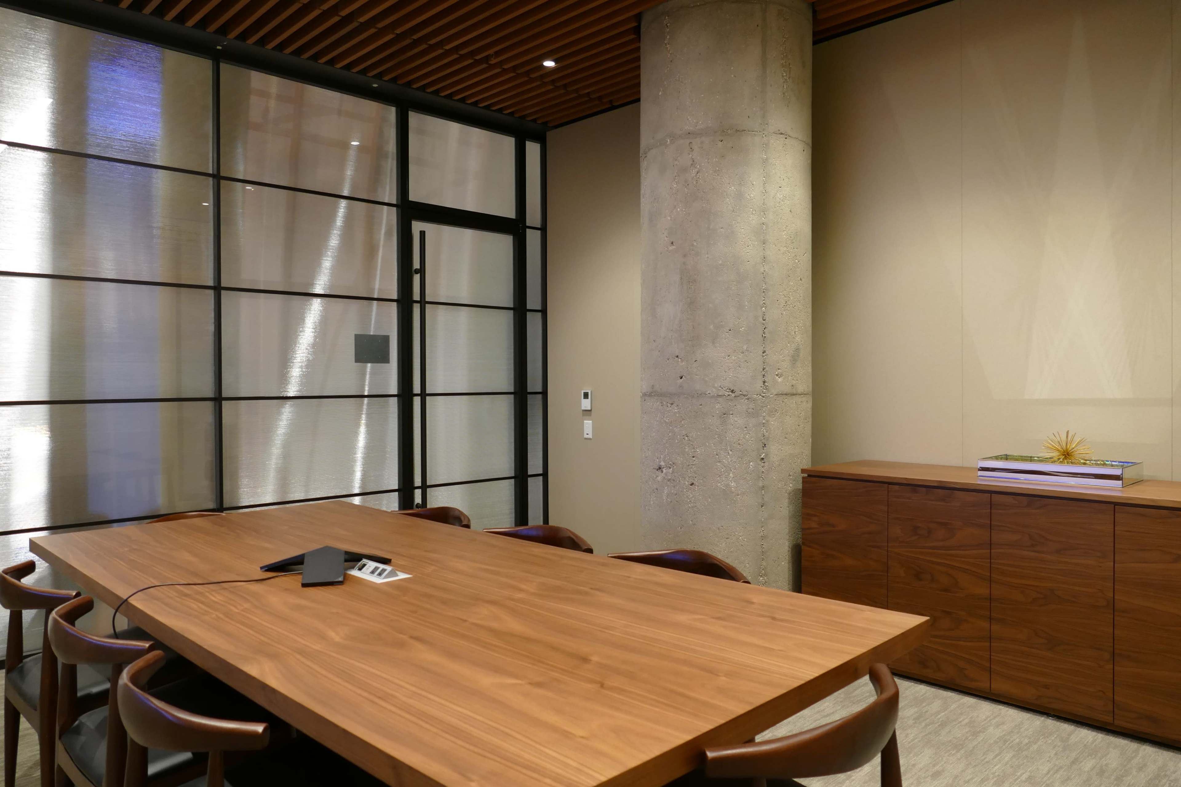 A modern meeting room features a wooden table surrounded by chairs, a concrete pillar, and a glass wall leading to another space.