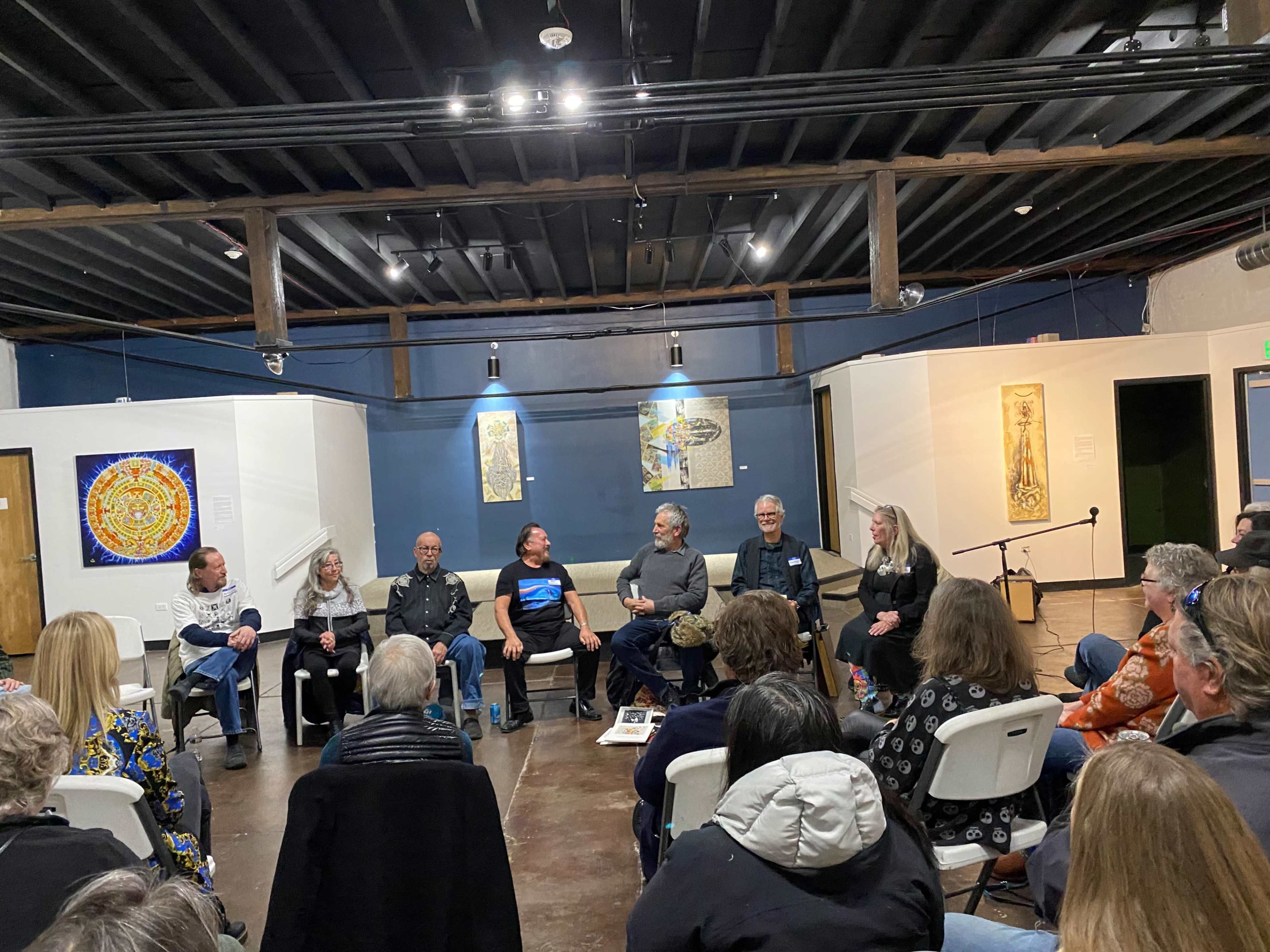 A group of eight people sits on chairs arranged in a semicircle in a spacious room with artwork on the walls, while an audience listens attentively.