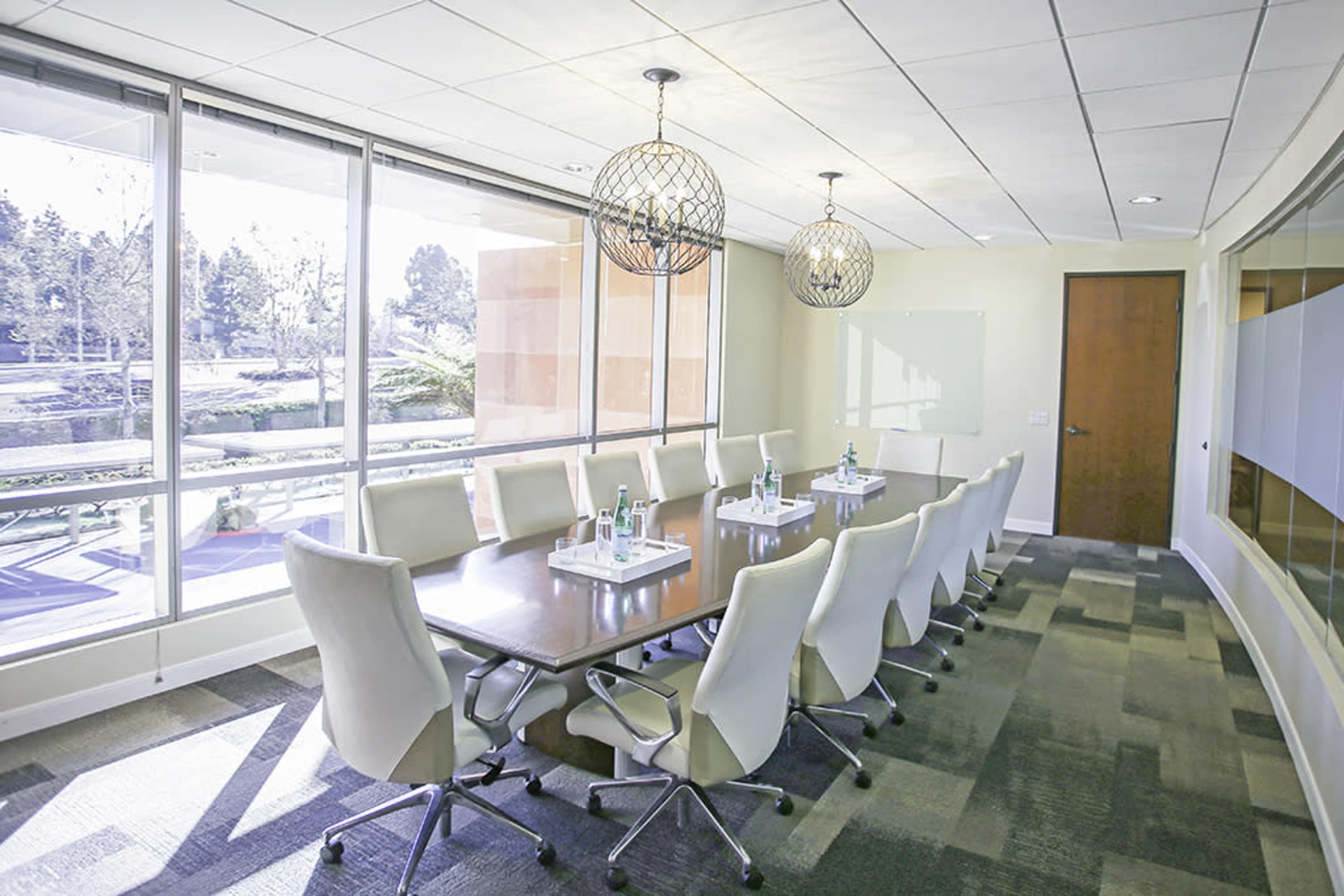 The image shows a modern conference room with a long table surrounded by chairs, large windows allowing natural light, and decorative light fixtures hanging above.