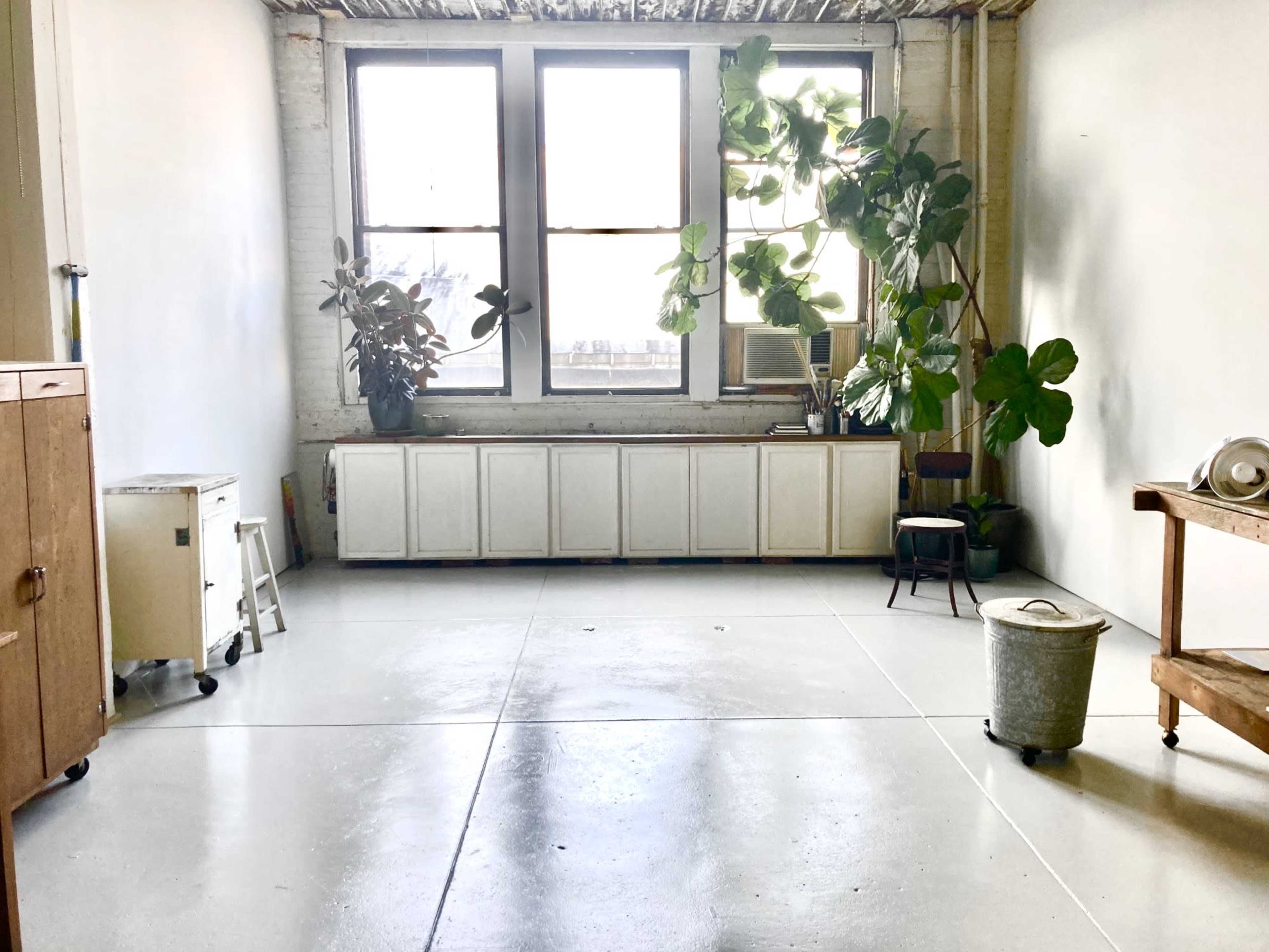 The image features a spacious, minimalistic room with large windows, potted plants, and simple furniture arrangements.