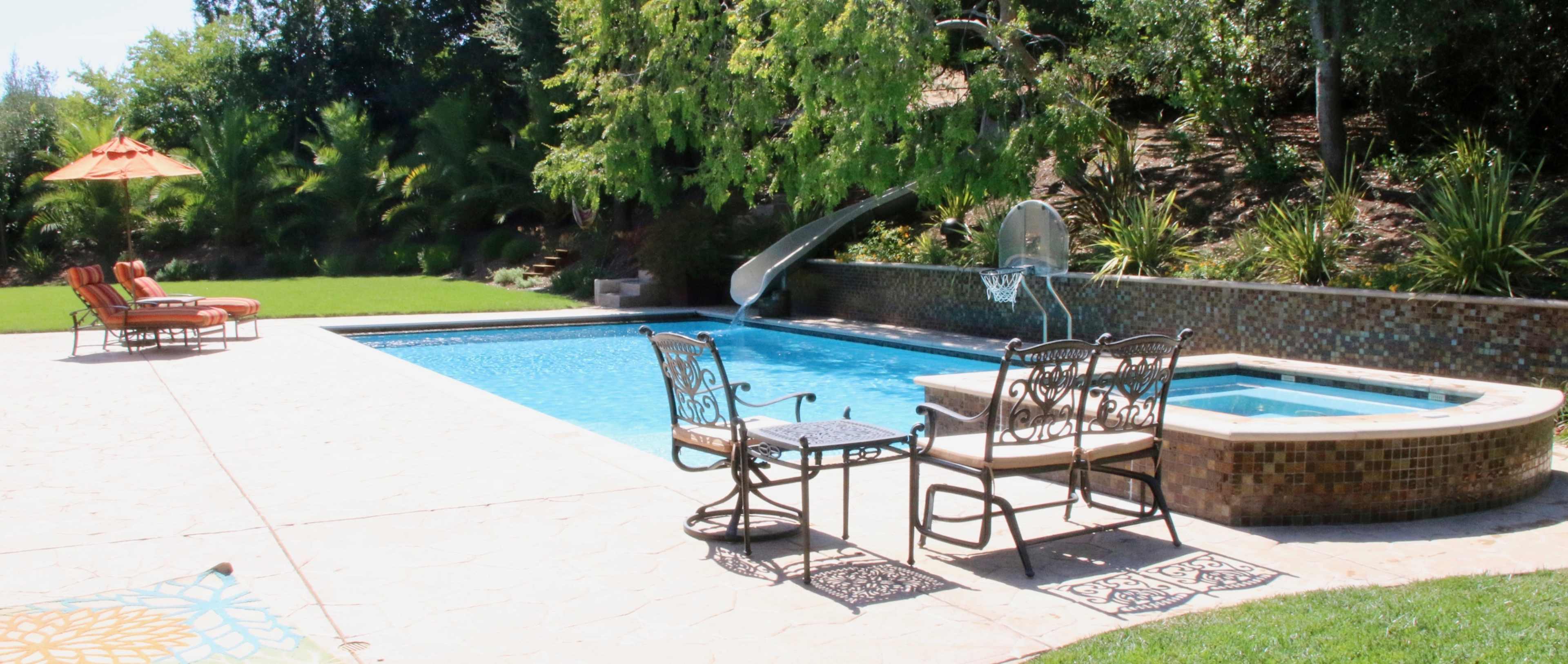 A backyard pool area with a slide, a hot tub, two lounge chairs, and a patio table surrounded by lush greenery.