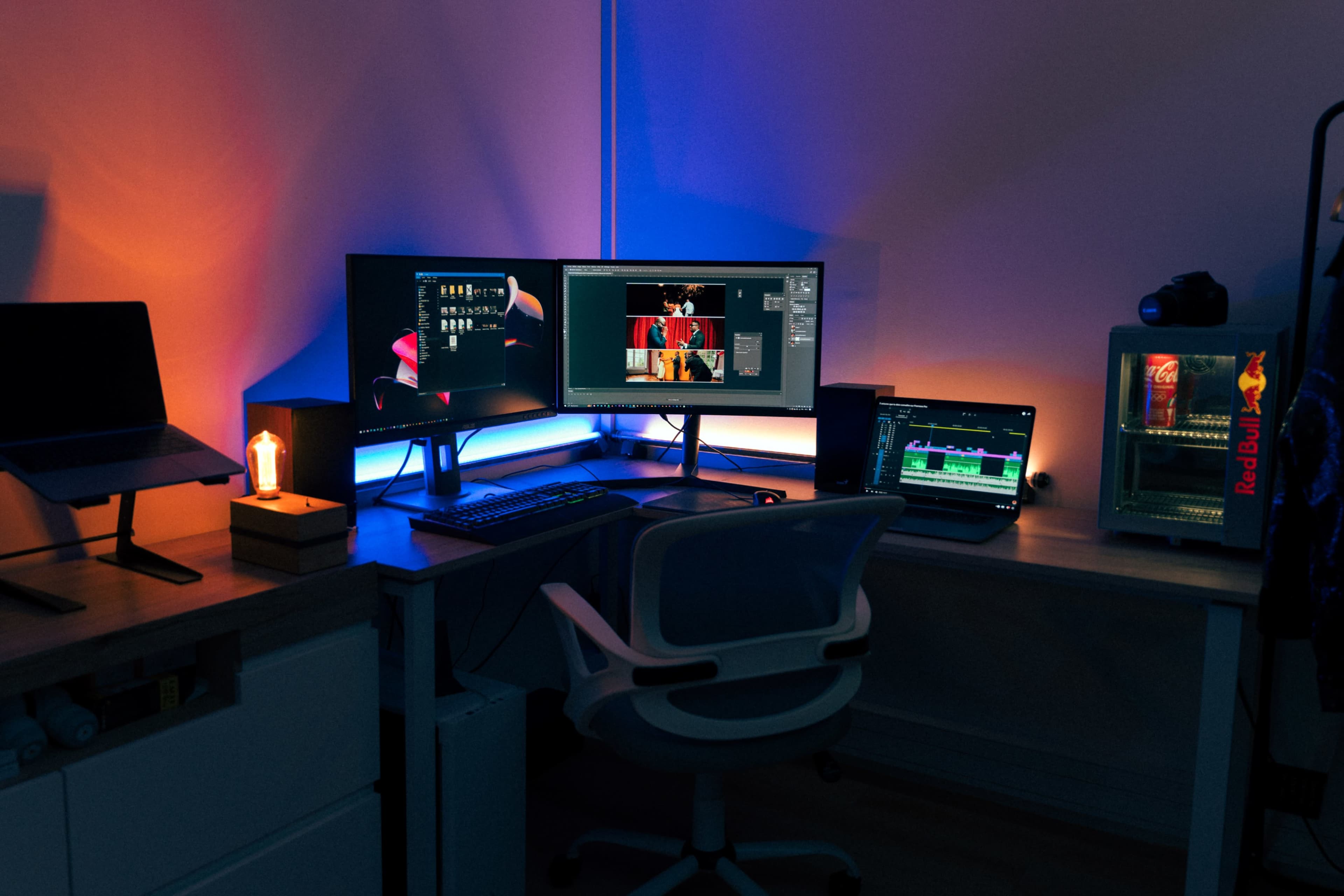 A workspace with two monitors set up for editing, a laptop on a lower surface, a desk lamp, and a refrigerator filled with drinks in a dimly lit room.
