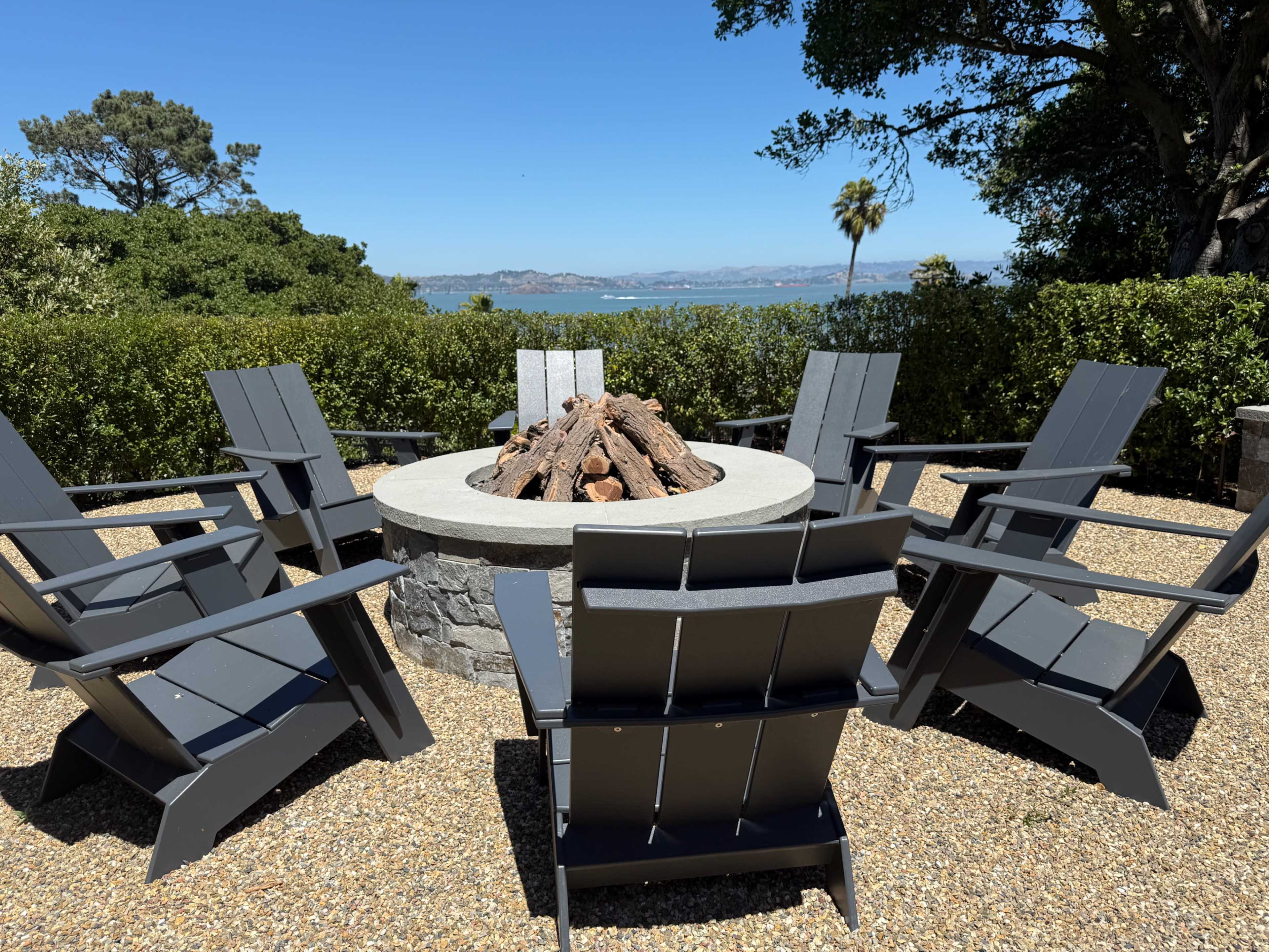 A circular stone fire pit surrounded by six gray Adirondack chairs on a gravel patio, with a view of greenery and a body of water in the background.