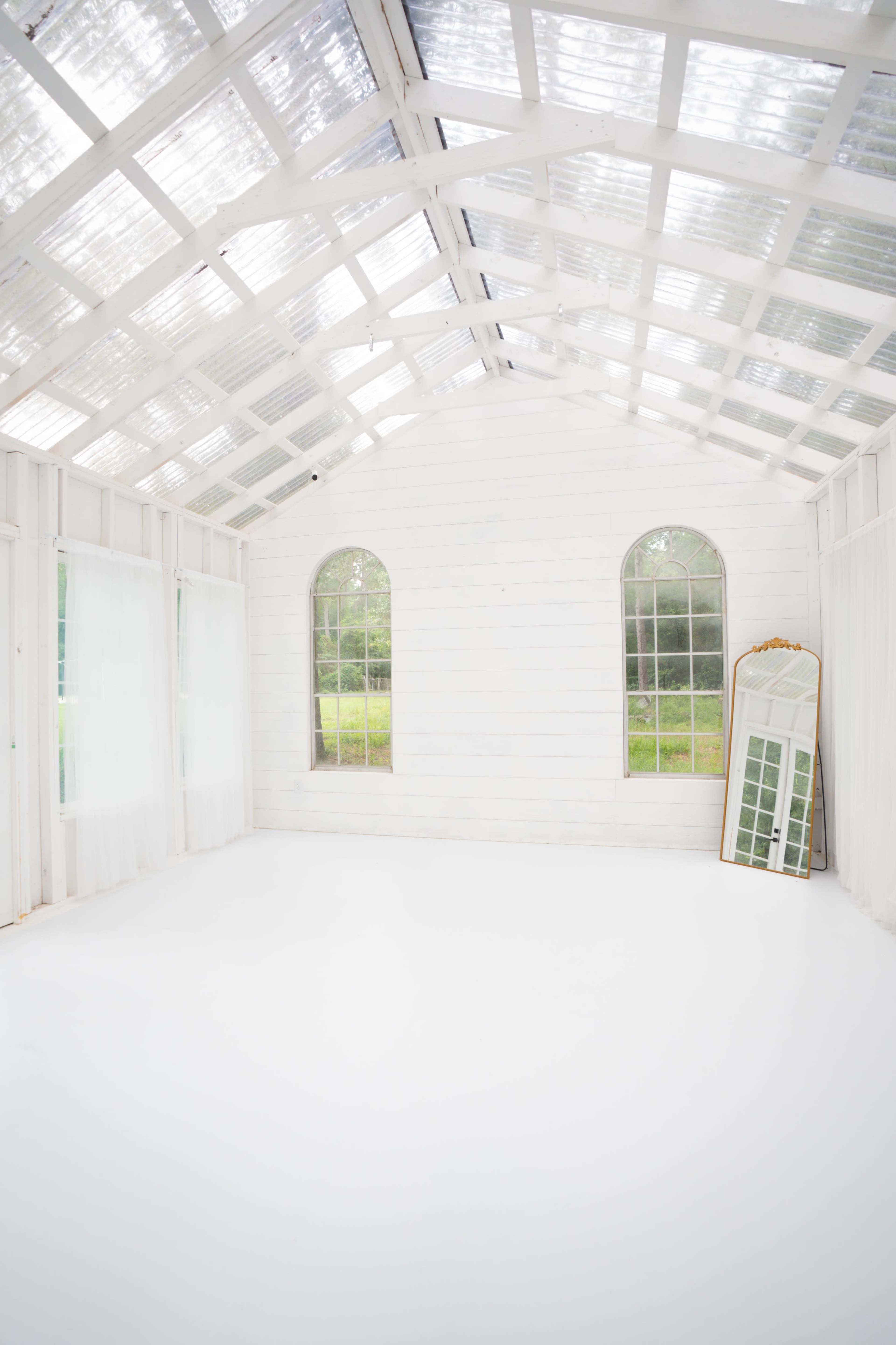 The image shows a bright, minimalist room with white walls, large arched windows, and a tall mirror leaning against the wall.