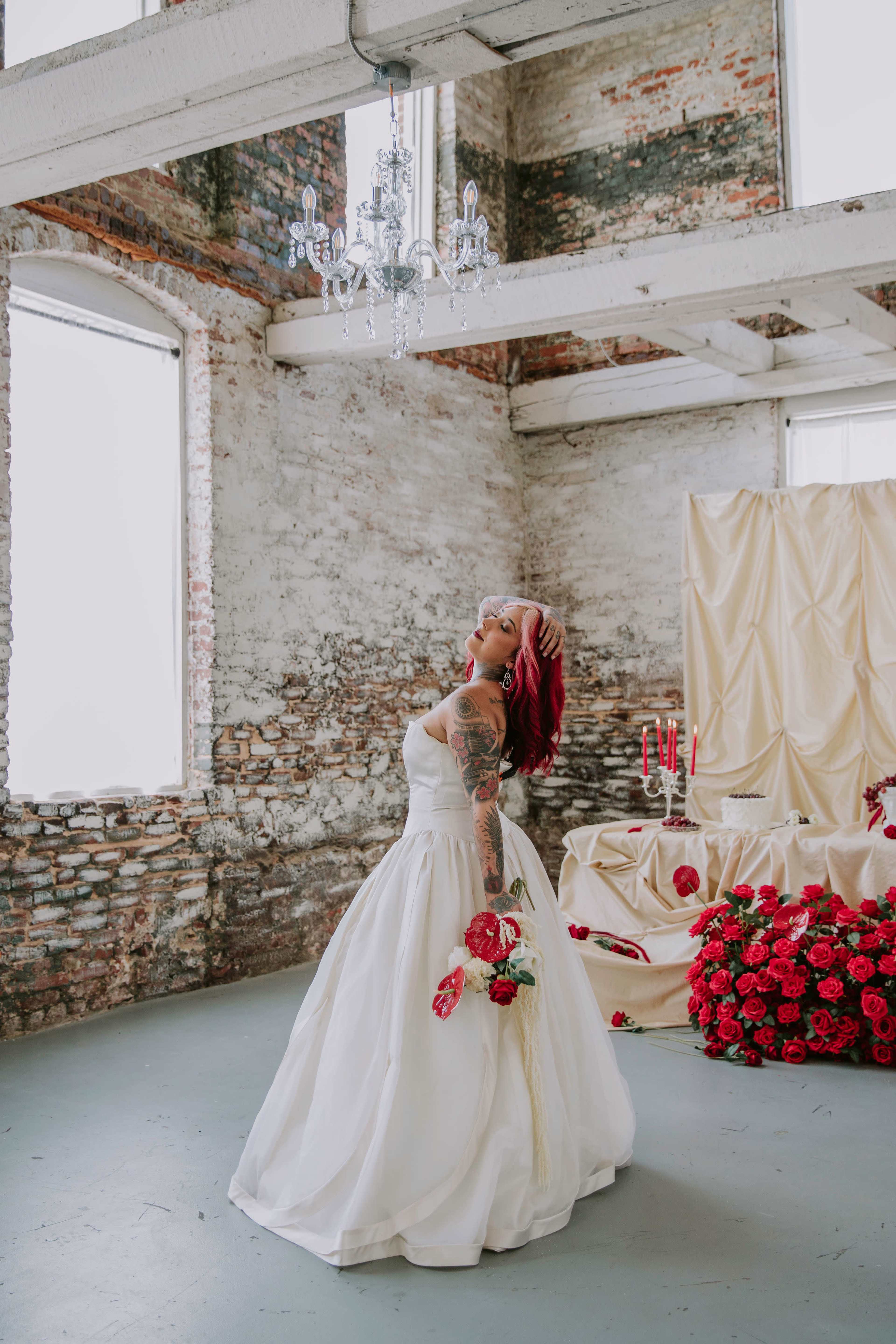 A woman in a white wedding dress stands in a rustic room with exposed brick walls, holding a bouquet of red flowers, while a decorated table with red roses and candles is nearby.
