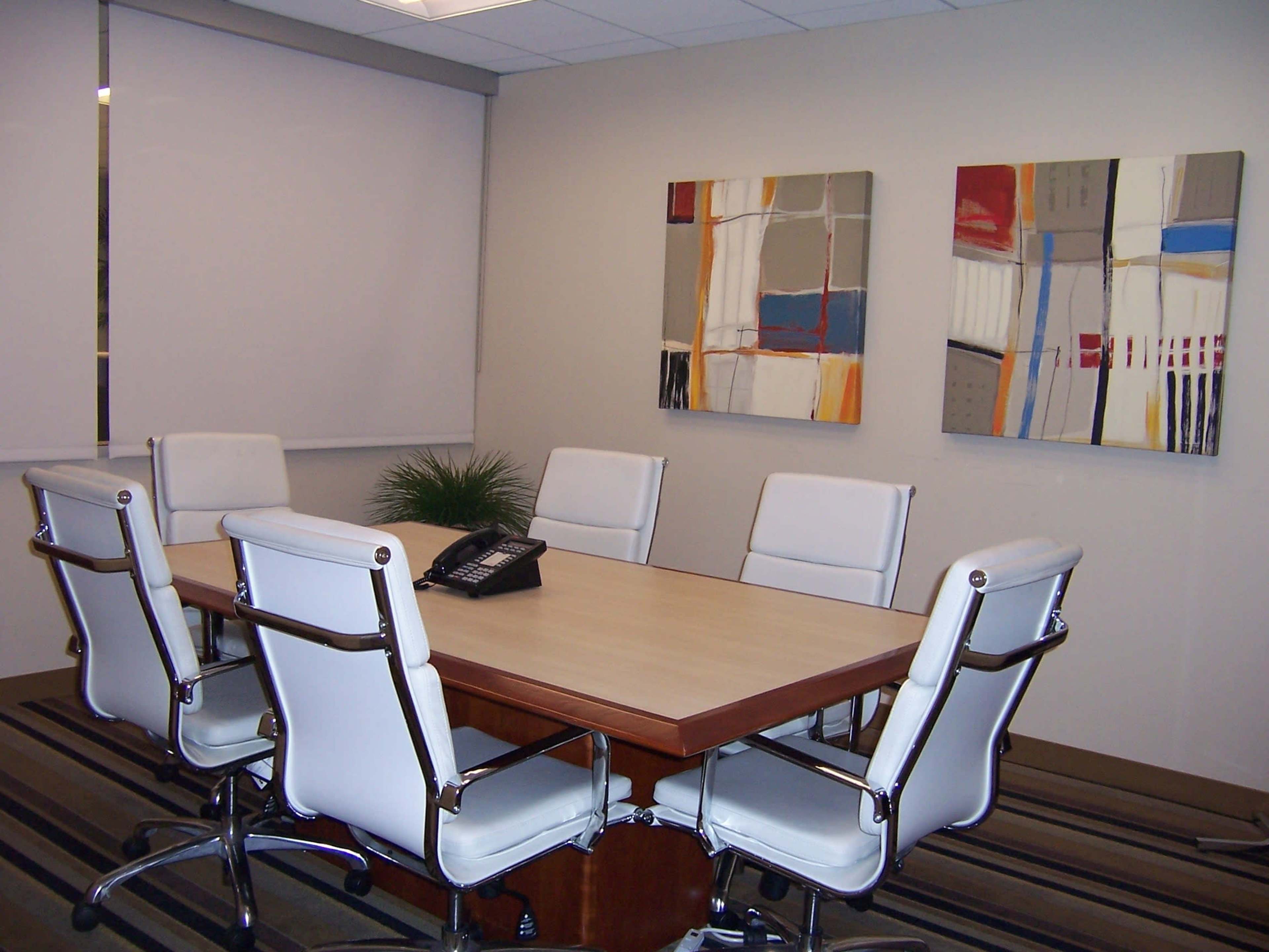 A conference room is set up with a rectangular table surrounded by six white chairs, and two abstract paintings are mounted on the wall.