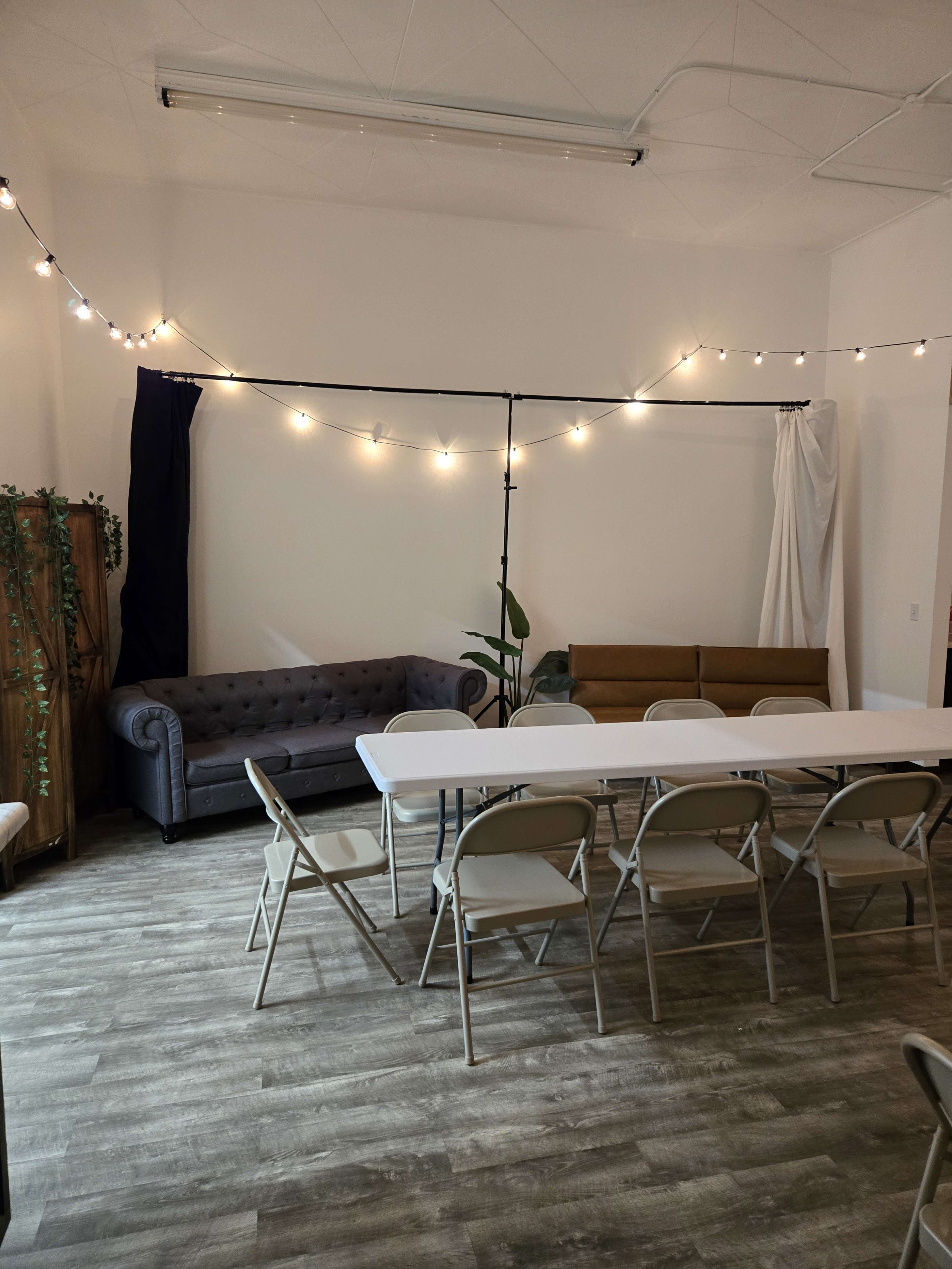 The image shows a well-lit room with a table set for several chairs, a blue couch, and a wooden bench against the walls, along with string lights overhead.