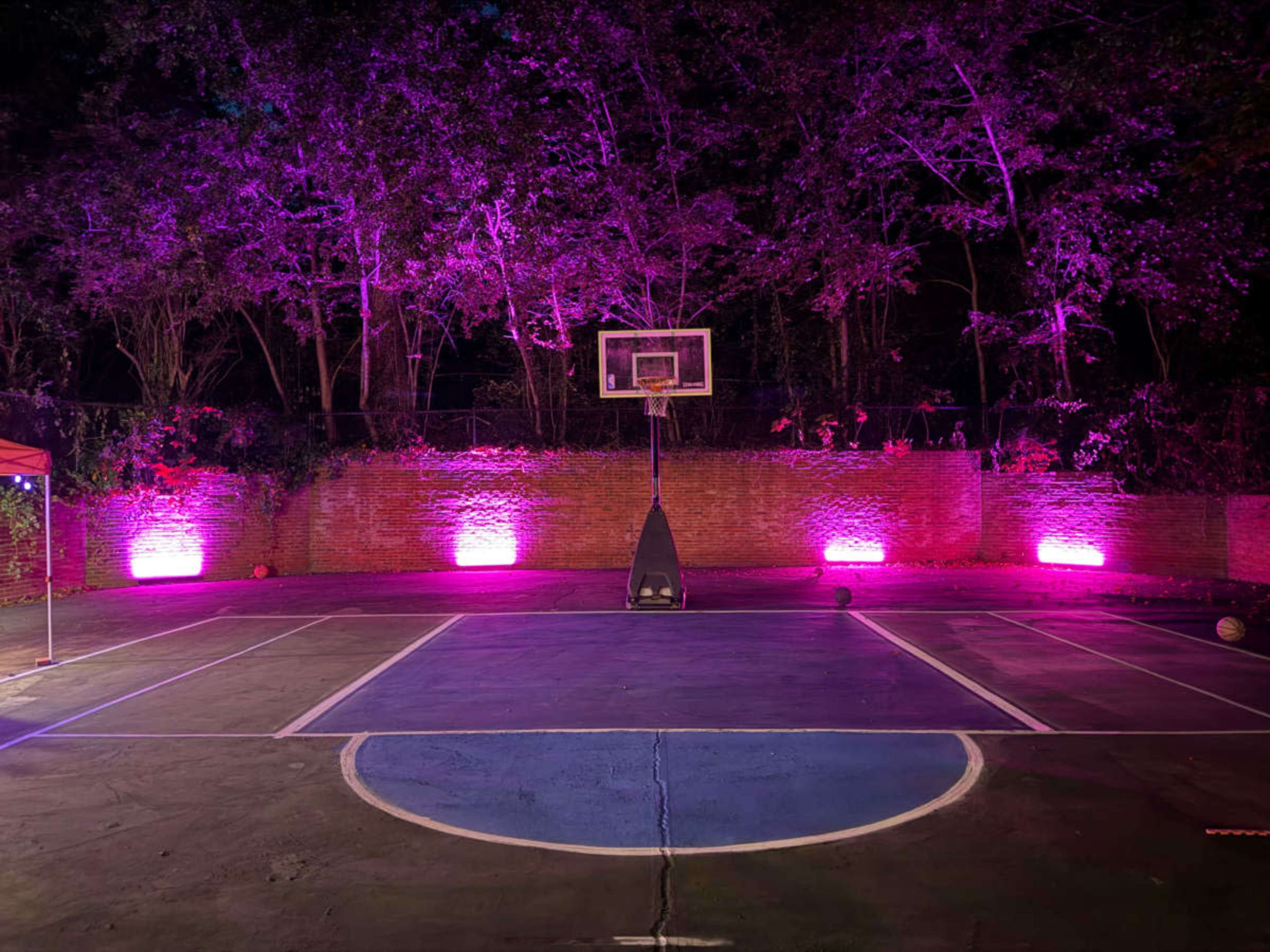 A basketball court is illuminated by purple lights, with a hoop at one end and a few basketballs scattered on the ground.
