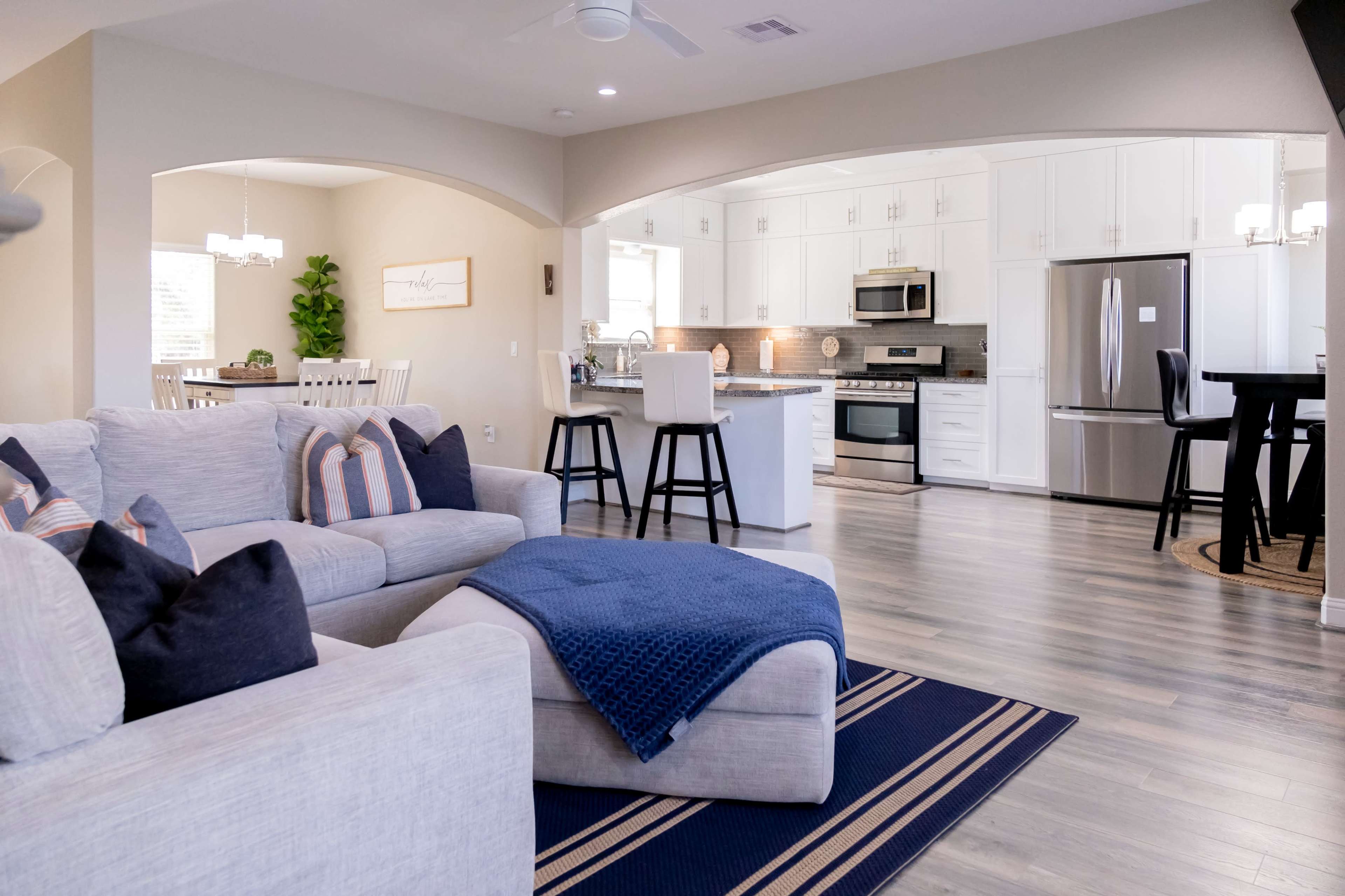 A modern open-concept living area with a beige sectional sofa, a blue throw blanket, and a kitchen featuring white cabinets and stainless steel appliances.