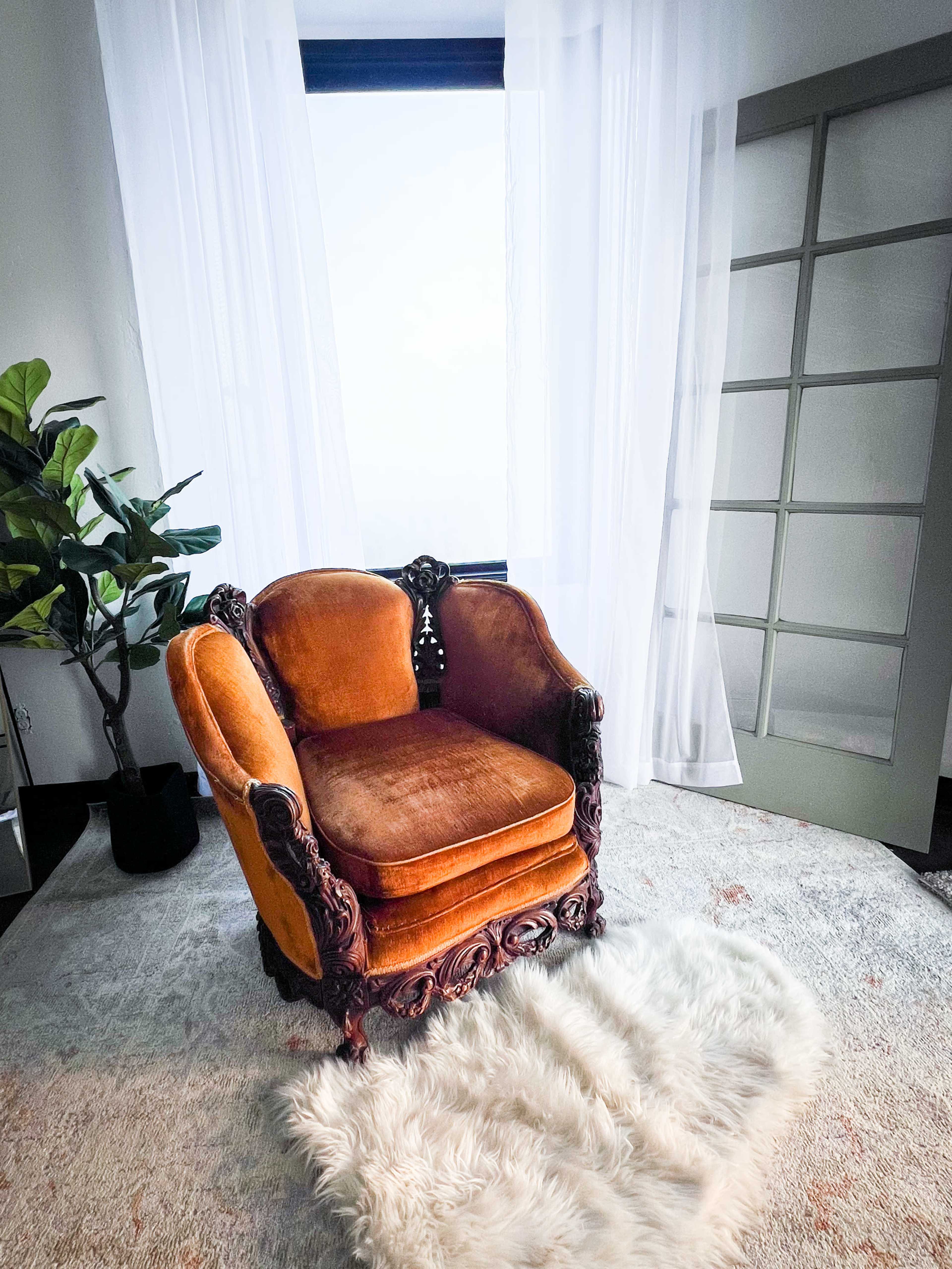 A vintage orange upholstered chair with intricate carvings sits on a fluffy rug in front of a large window draped with sheer curtains.