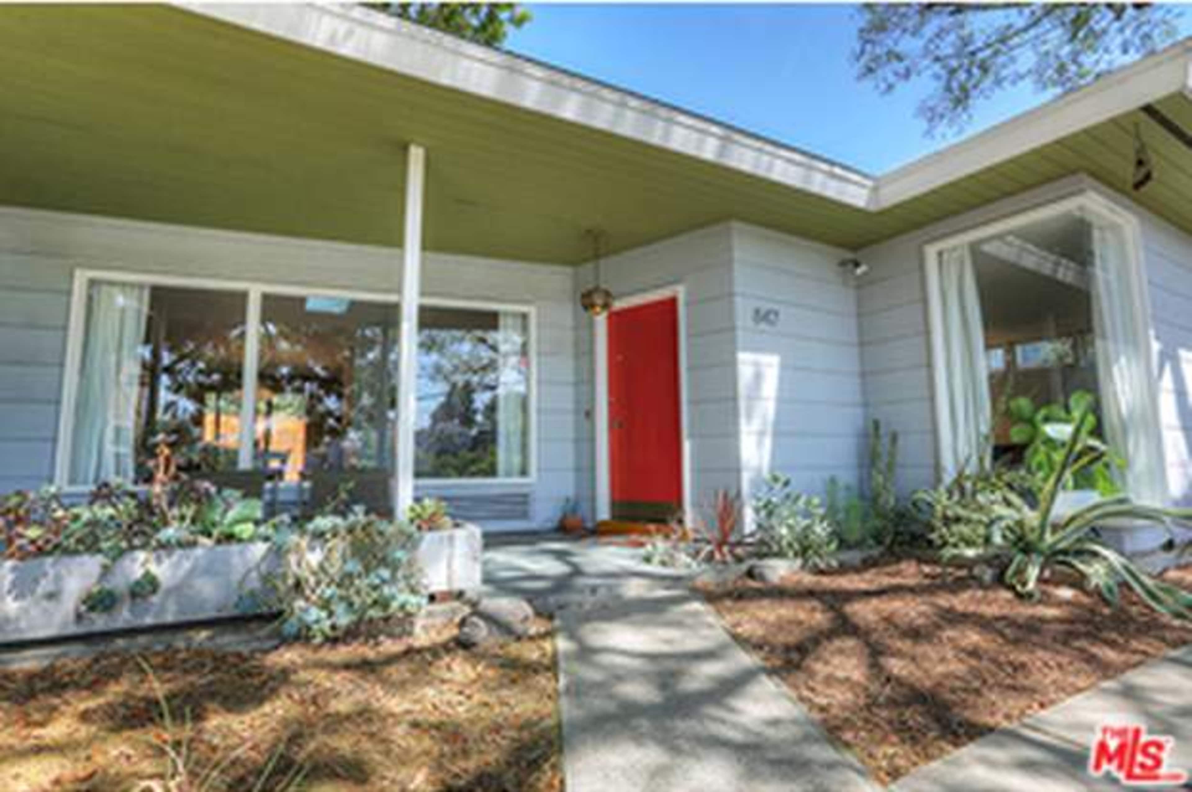 A modern single-story house with a red front door and large windows is surrounded by landscaped shrubs and pathways.