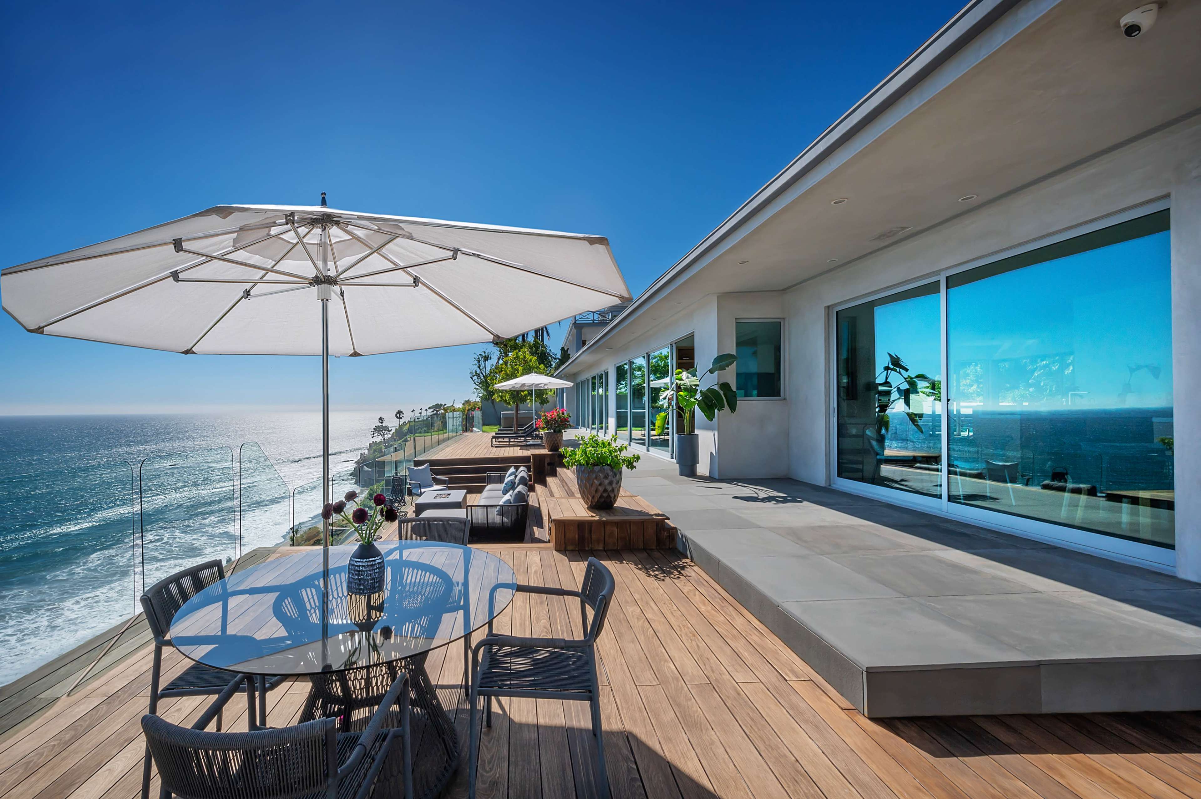 A modern terrace features a glass dining table and an umbrella, overlooking the ocean with lounge furniture and potted plants.