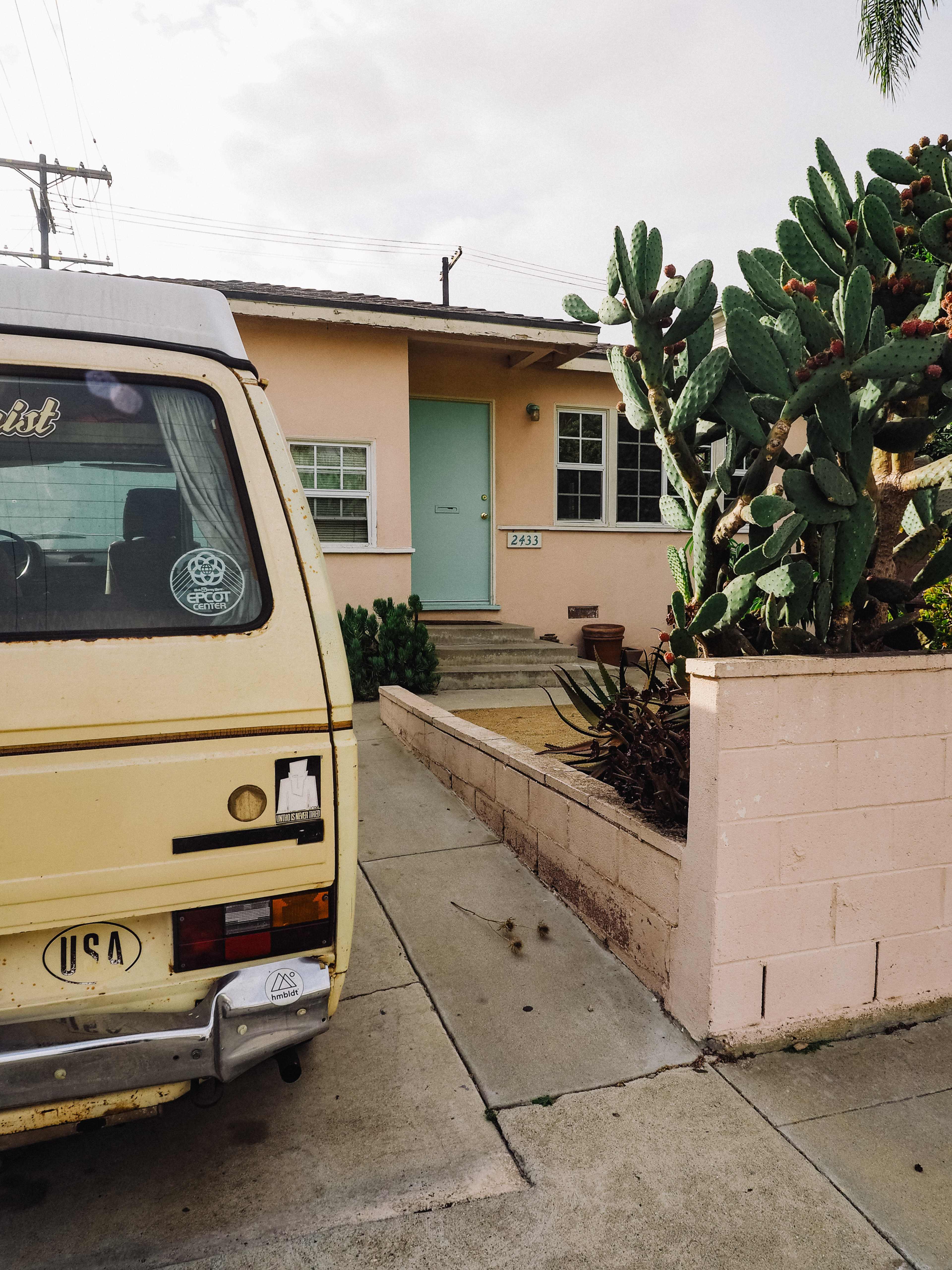 A yellow van is parked in front of a single-story pink house with a green door and a front yard featuring cacti.