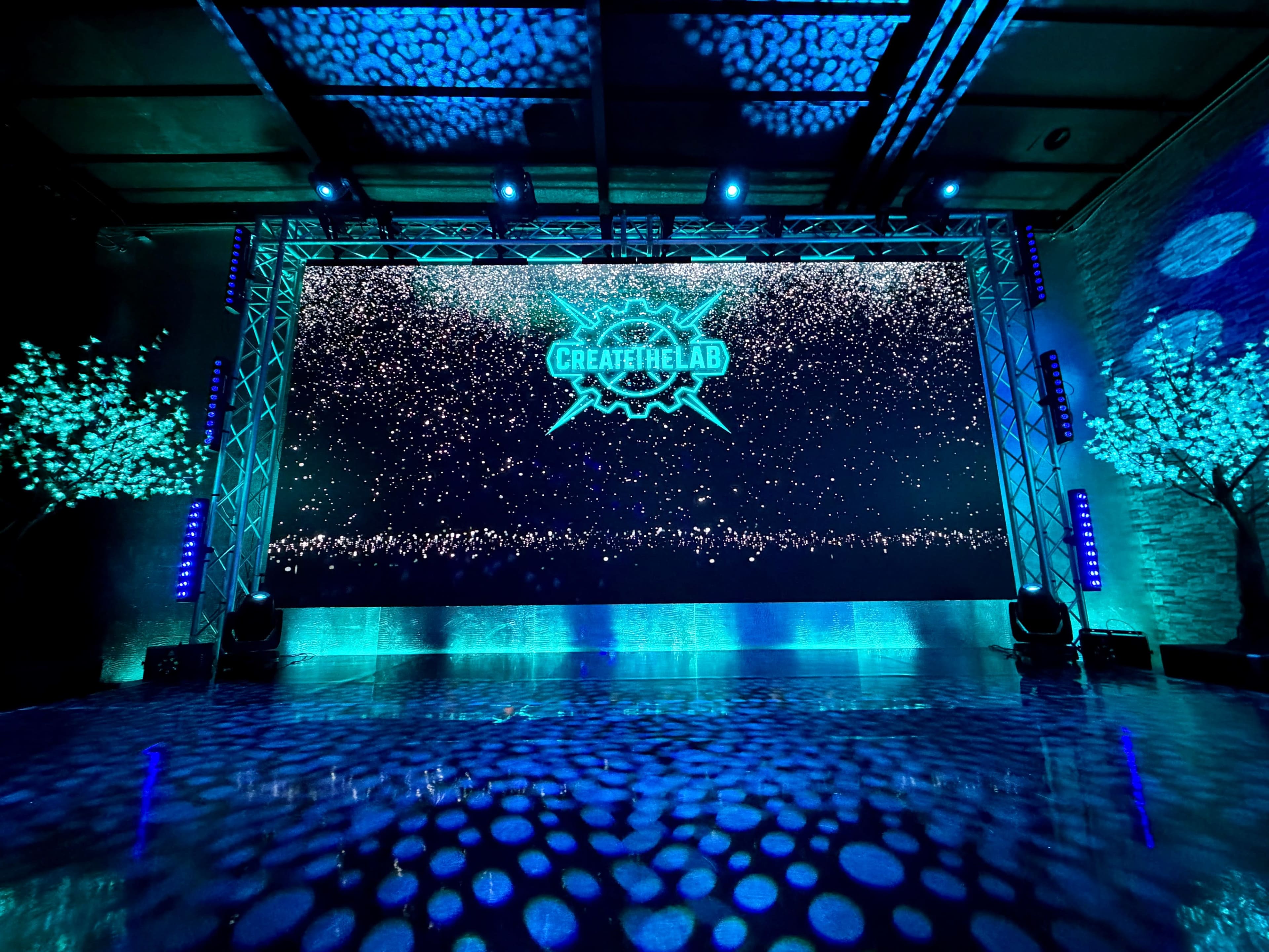 The scene features a large illuminated screen displaying the words "CREATETHELAB," surrounded by decorative trees and dynamic lighting effects in a darkened venue.