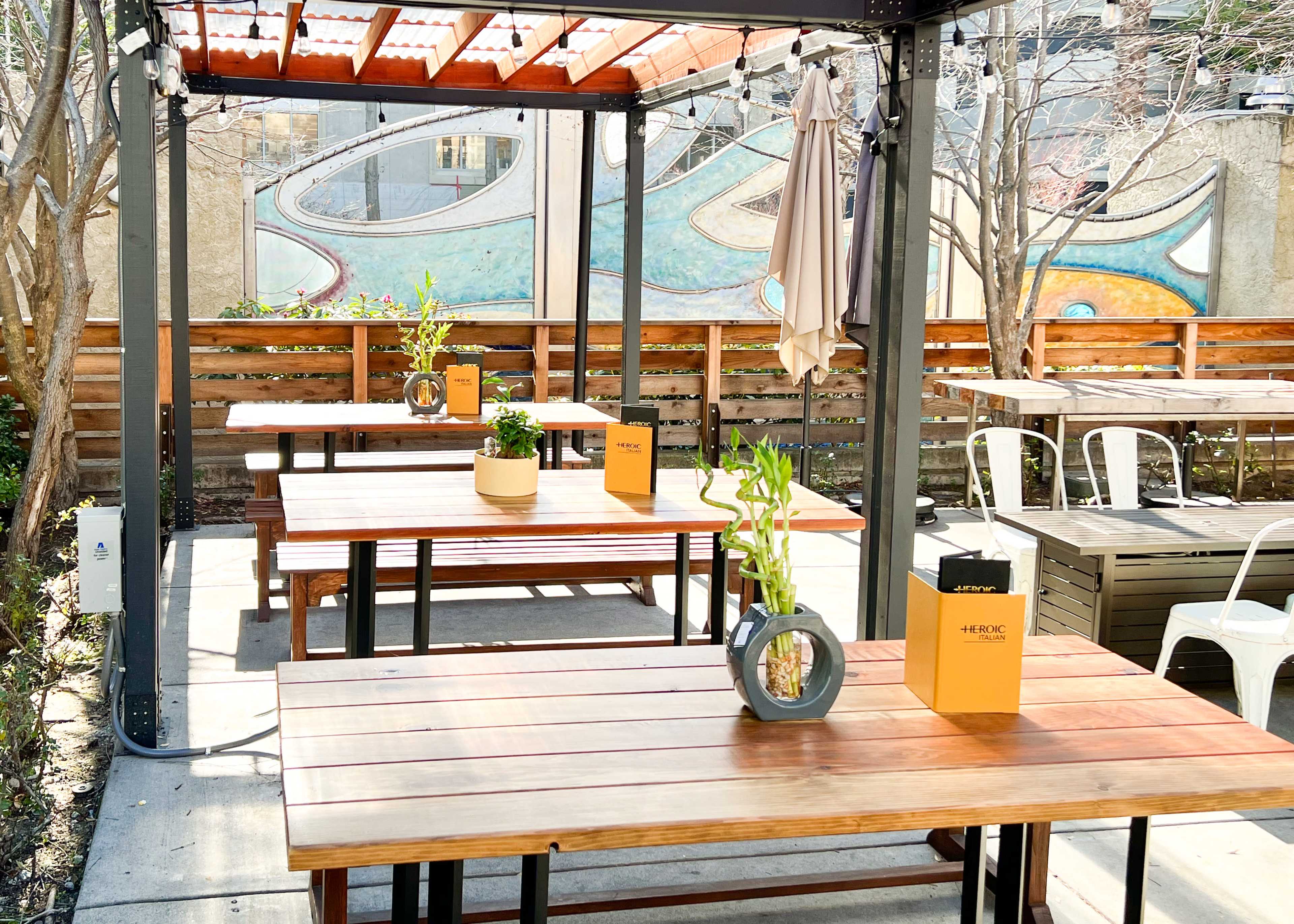 An outdoor dining area features wooden tables with potted plants and a colorful mural in the background.