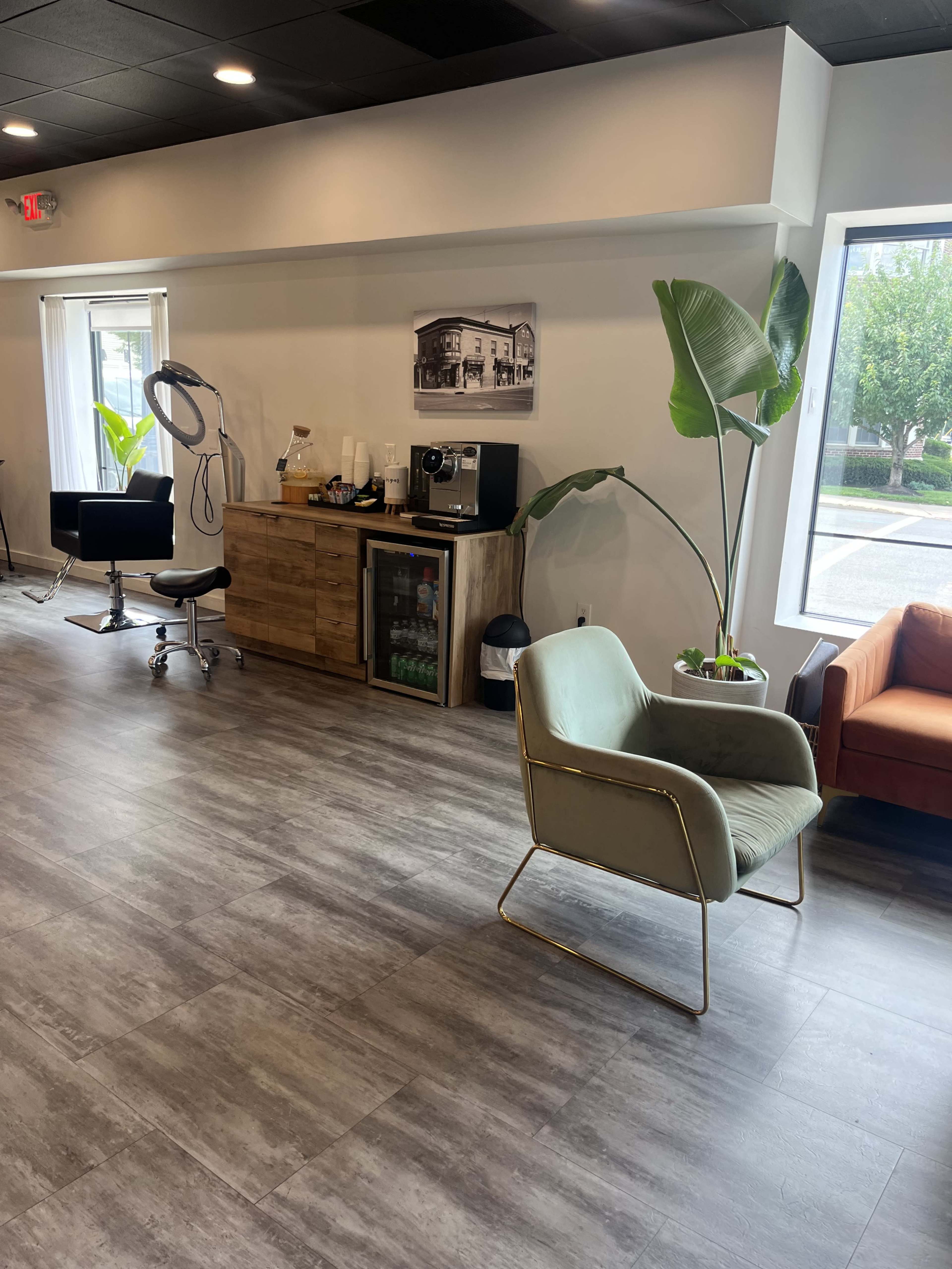 The image shows a modern interior space featuring a green chair, a wooden cabinet with a coffee machine and mini-fridge, a salon chair, and large windows with natural light.