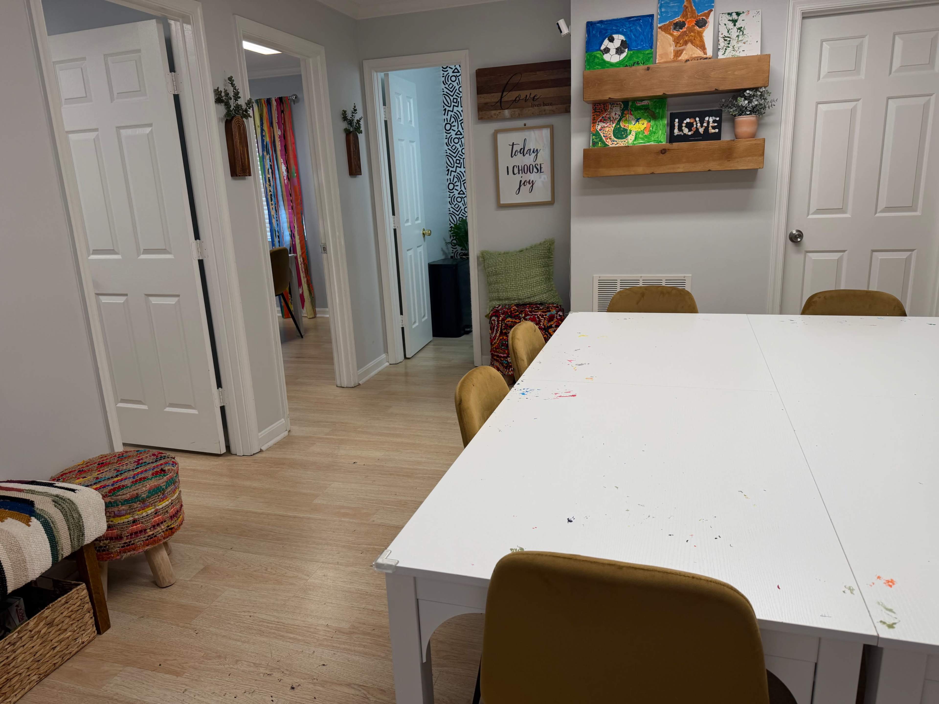 The image shows a bright, organized room with a large white table surrounded by yellow chairs, leading to multiple doorways and colorful artwork on the walls.
