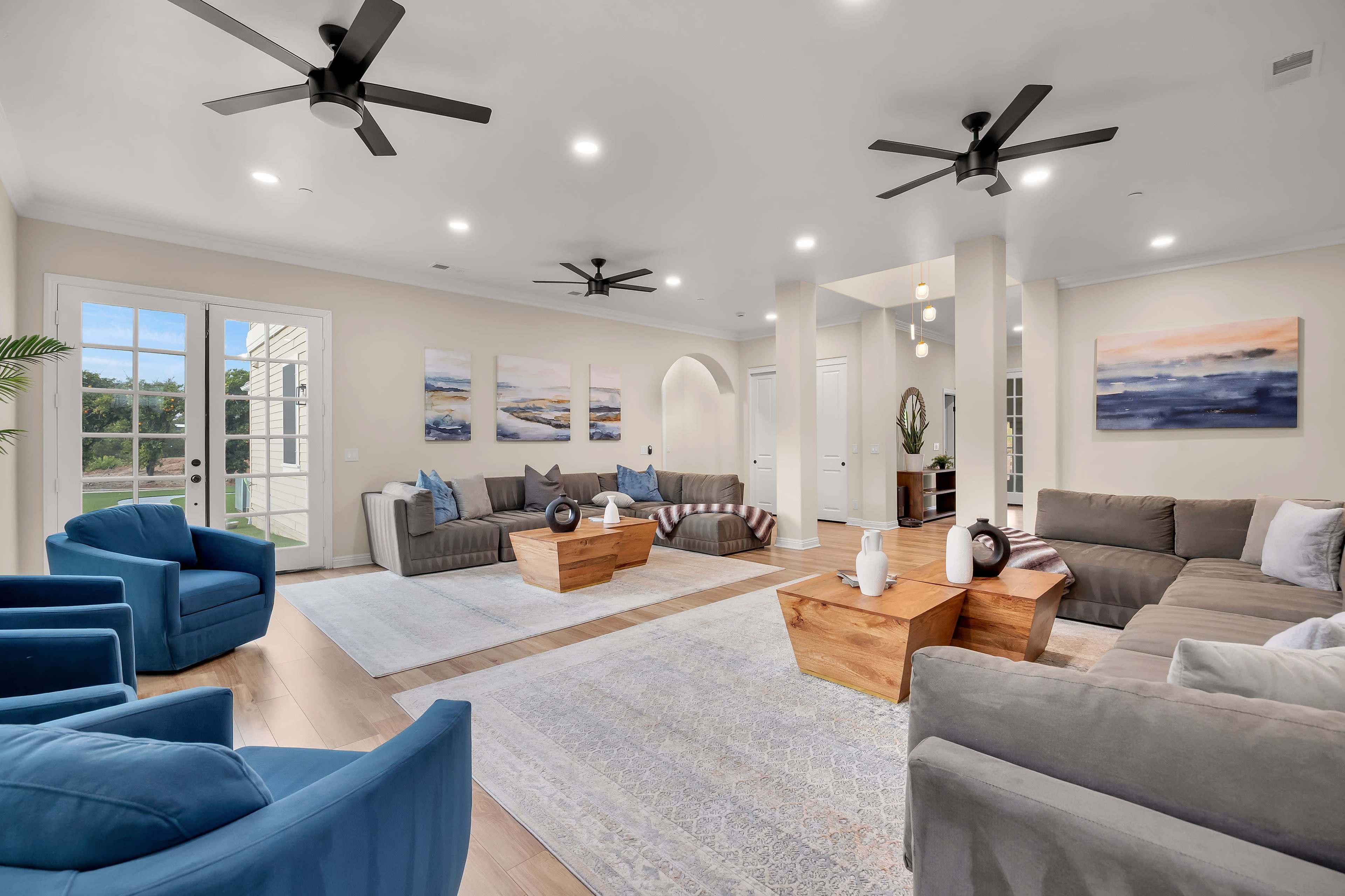 A spacious living room features two gray sectional sofas, blue accent chairs, wooden coffee tables, and large windows letting in natural light.