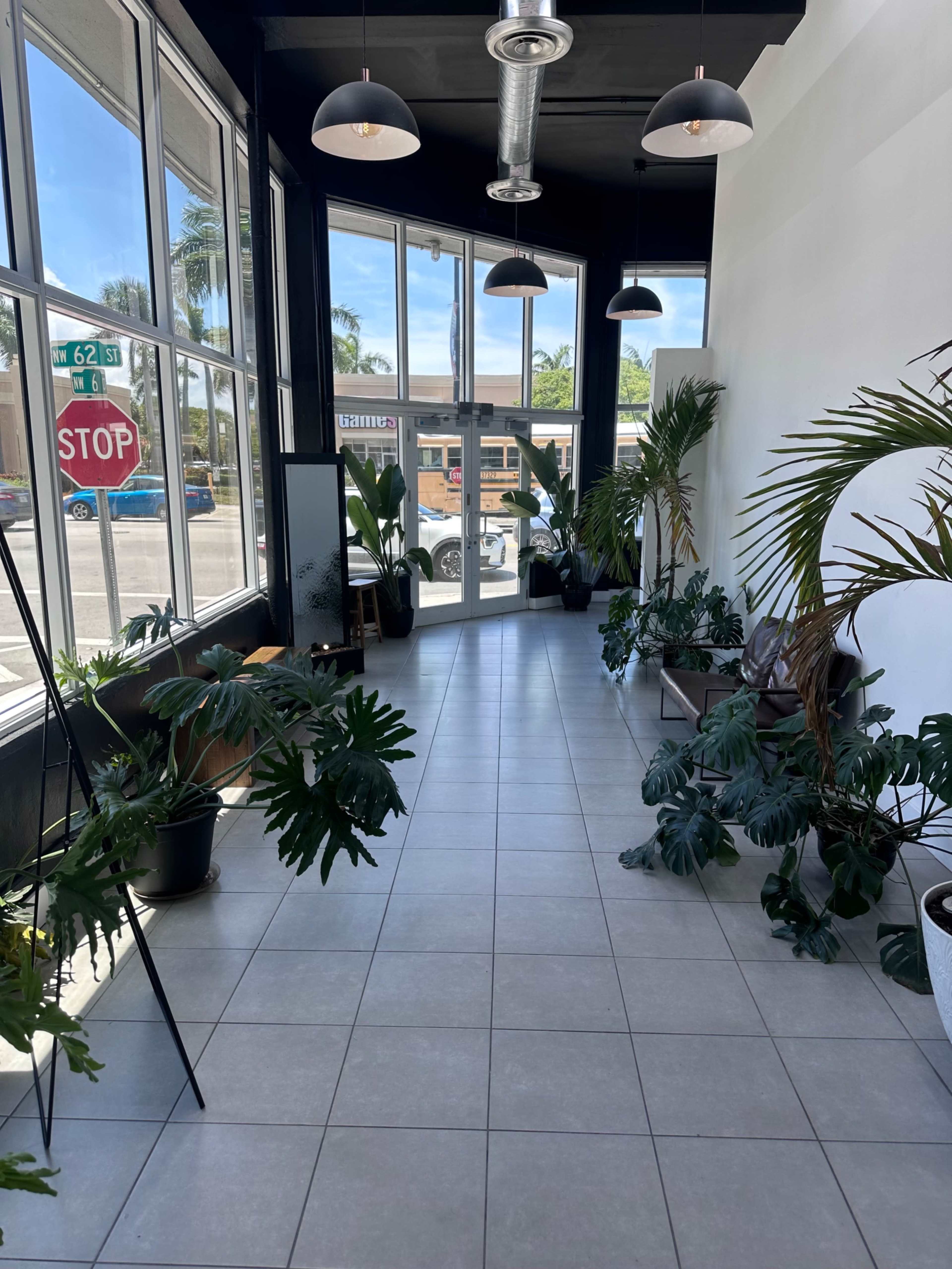 The image shows a spacious hallway with large windows, potted plants, and modern lighting fixtures.