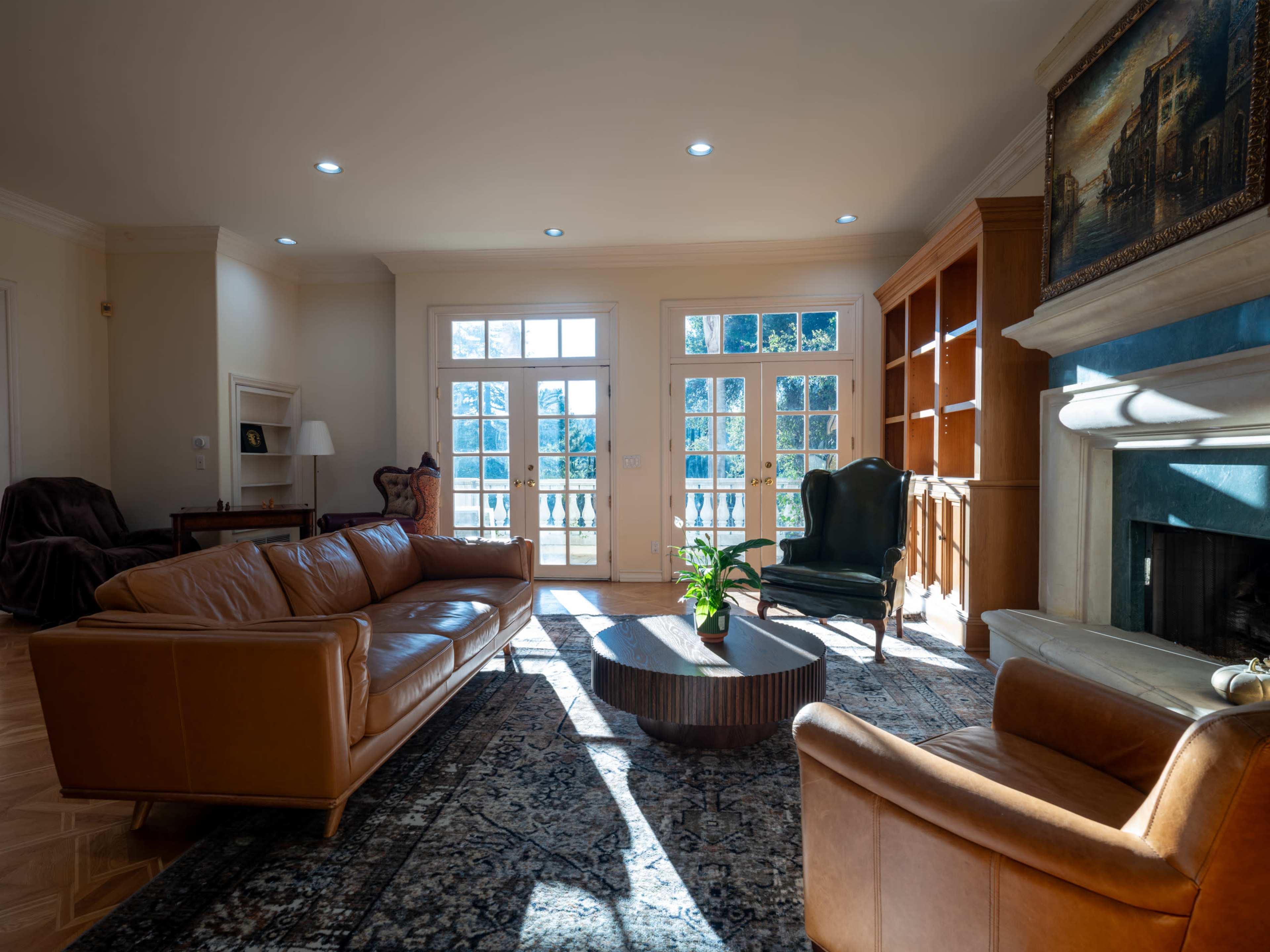 The image shows a well-lit living room featuring a brown leather sofa, a round wooden coffee table, a plush armchair, and large windows that open to a view.