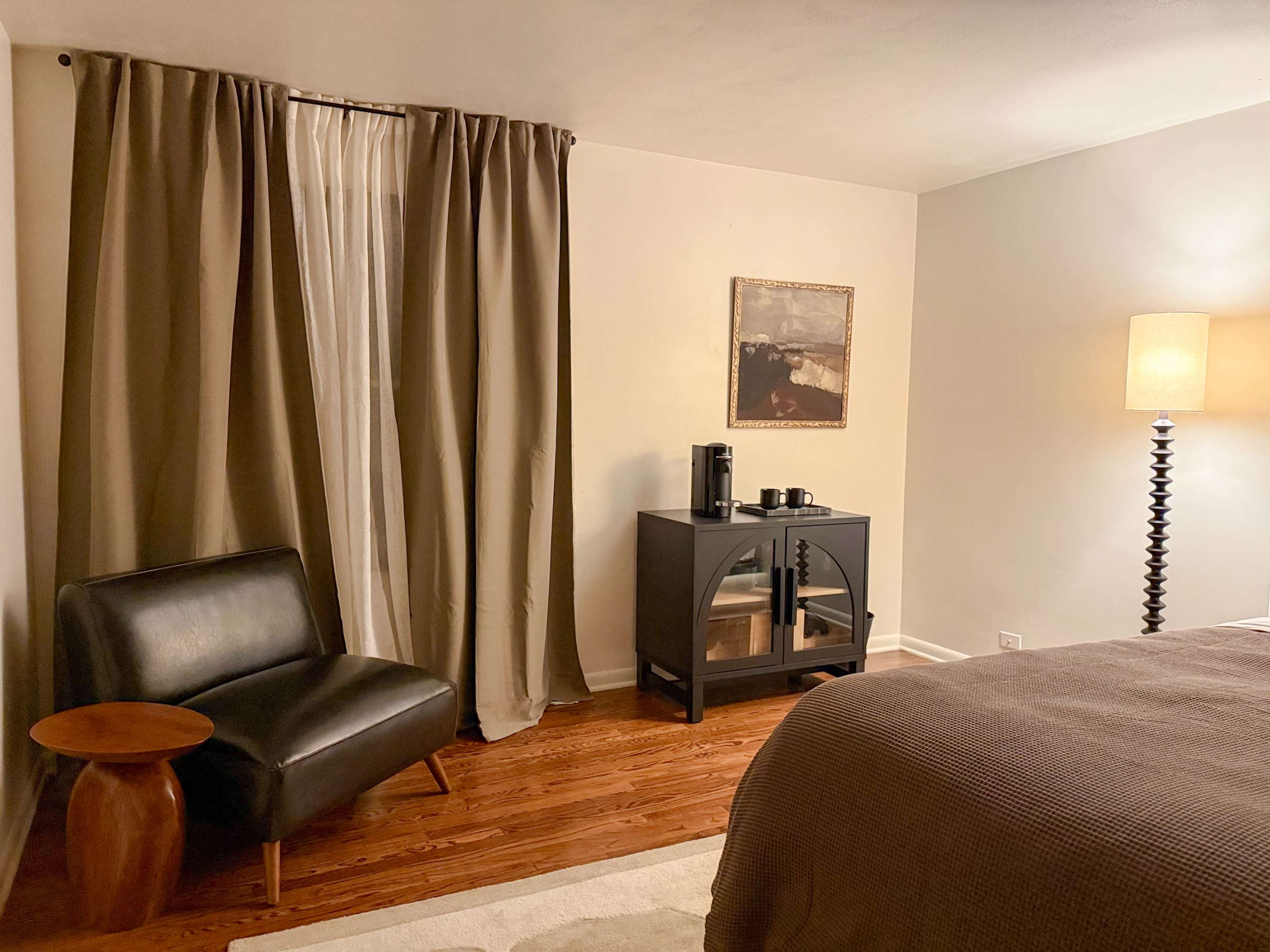 The image shows a cozy bedroom corner featuring a black upholstered chair, a round wooden side table, a dark cabinet, and a lamp next to a bed, with floor-length curtains framing a window.