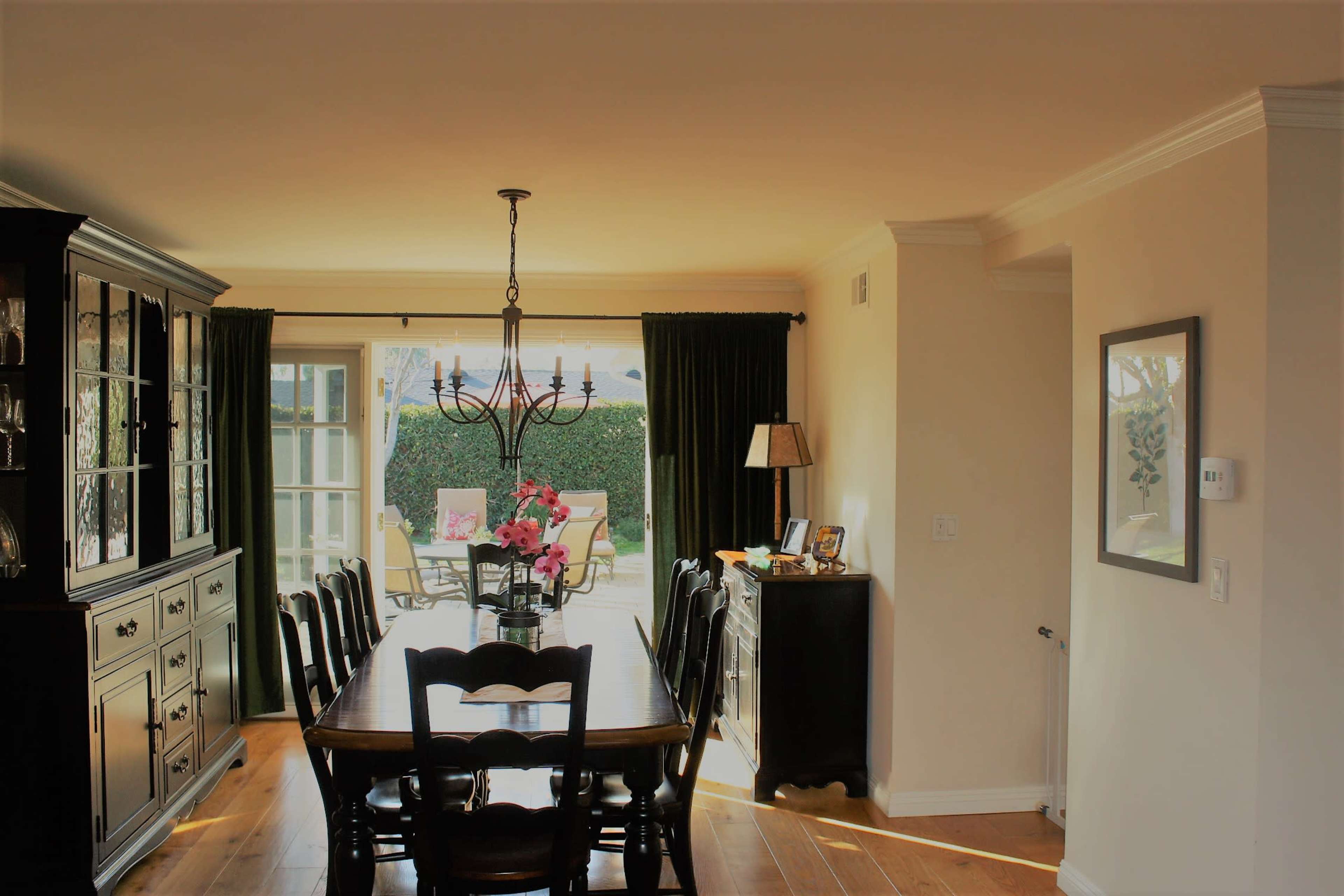 A dining room with a long wooden table and a chandelier, leading to a windowed area open to an outdoor space.
