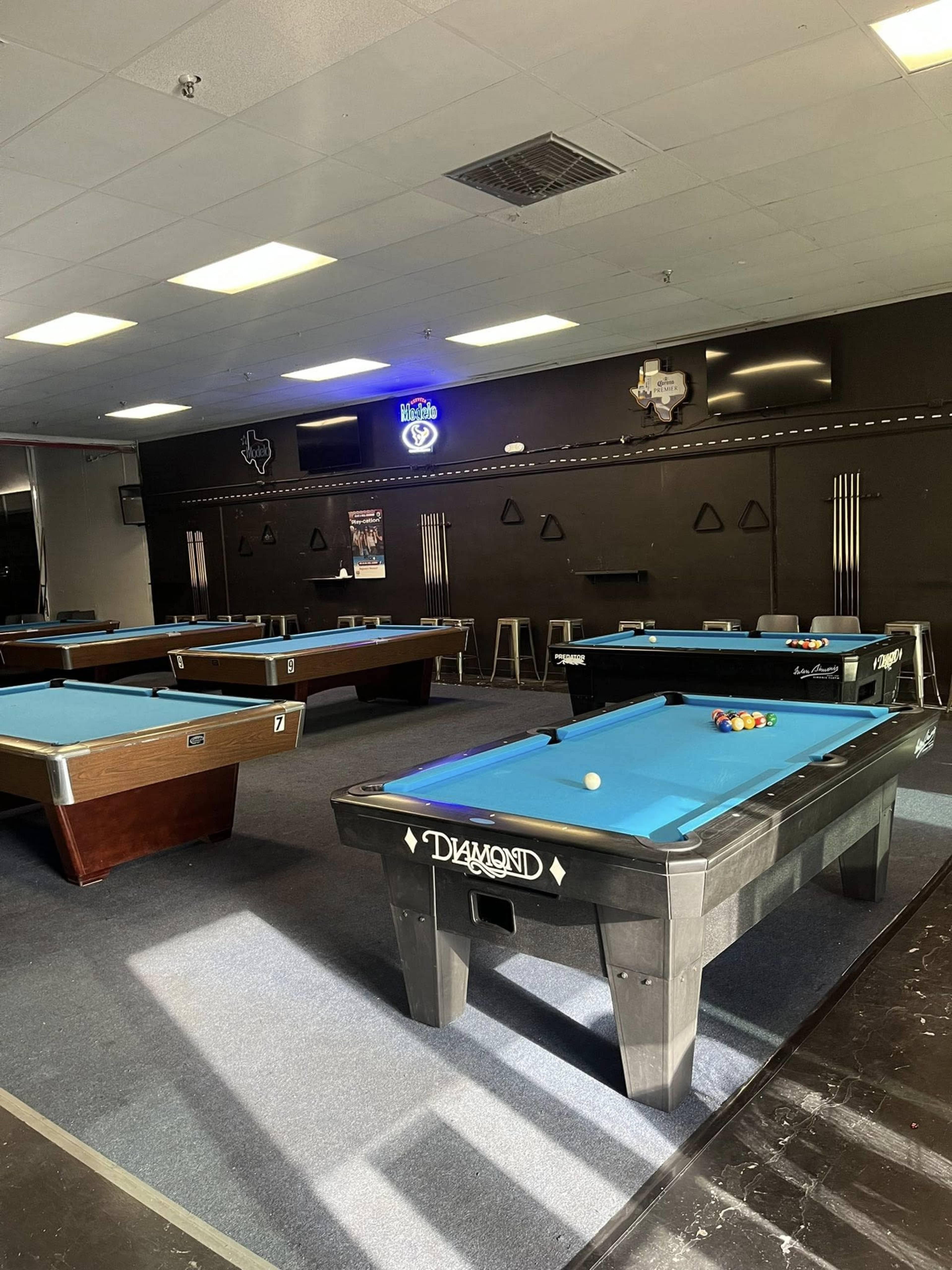 The image shows a spacious pool hall featuring multiple pool tables with blue felt surfaces arranged in a well-lit room.