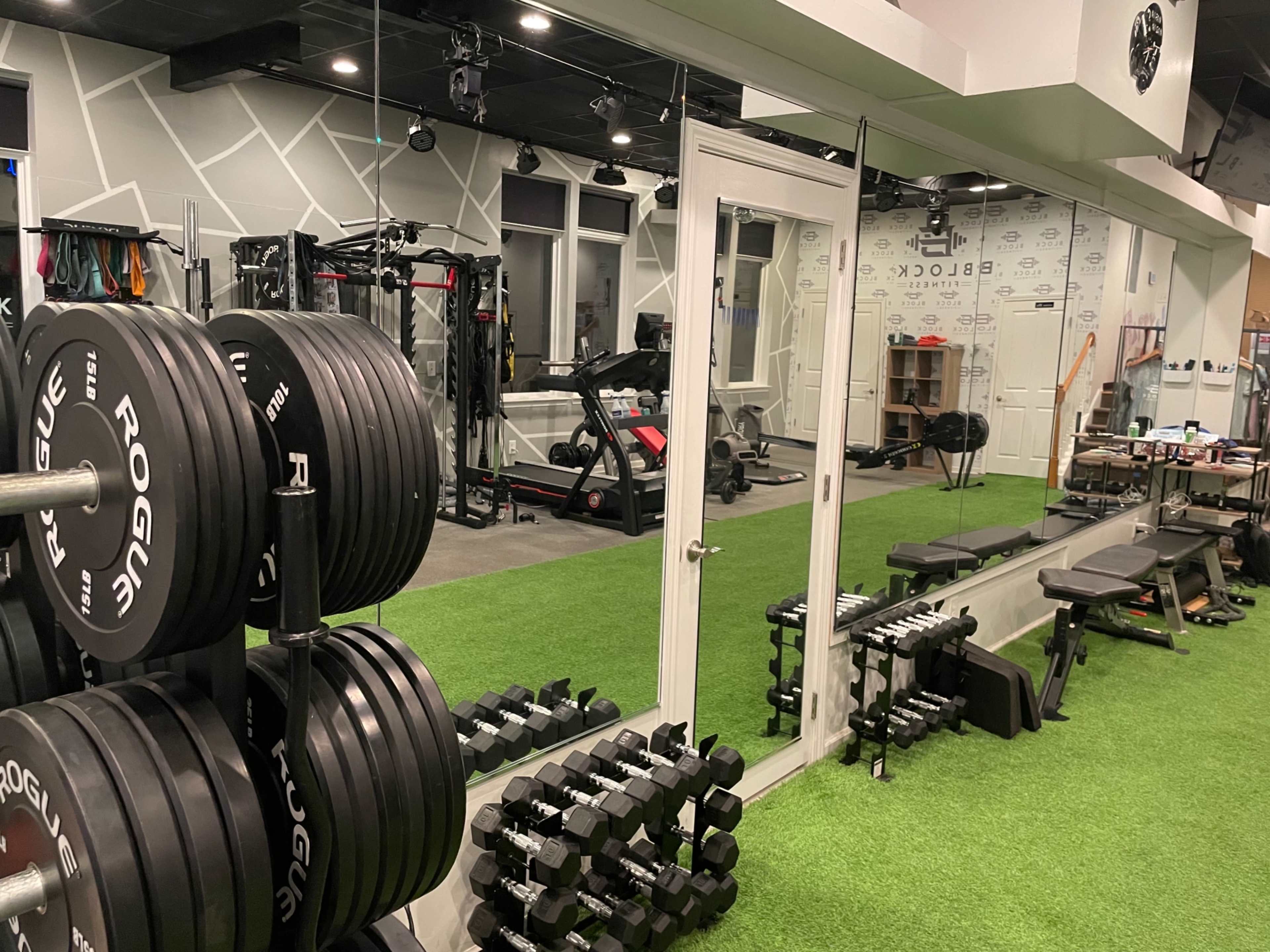 The image shows a modern home gym featuring weight plates, dumbbells, a bench, and a treadmill, all arranged alongside mirrored walls and artificial turf flooring.