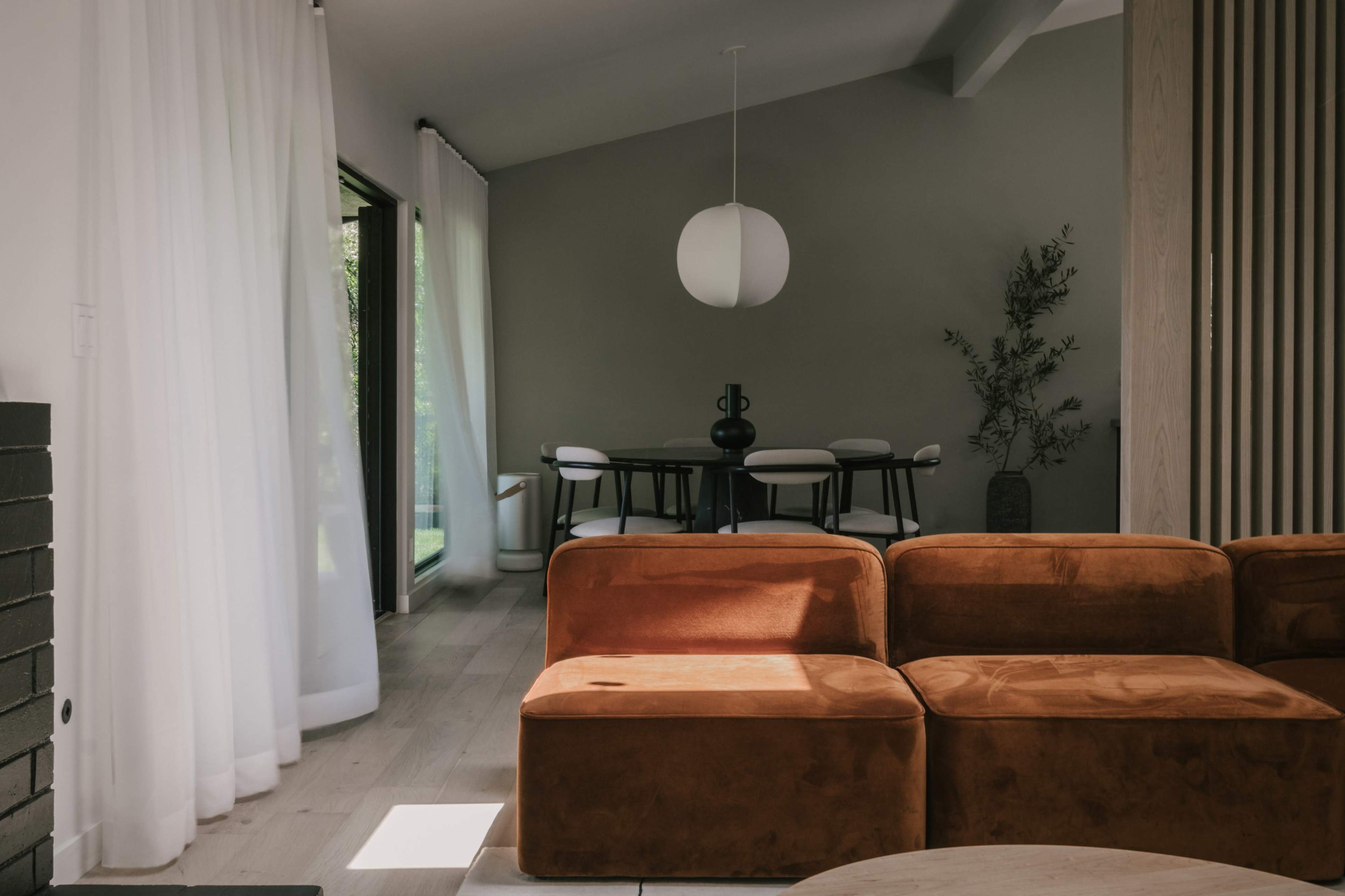 A modern living space featuring an orange sofa, a dining area with a round table and chairs, and large windows with sheer curtains letting in natural light.