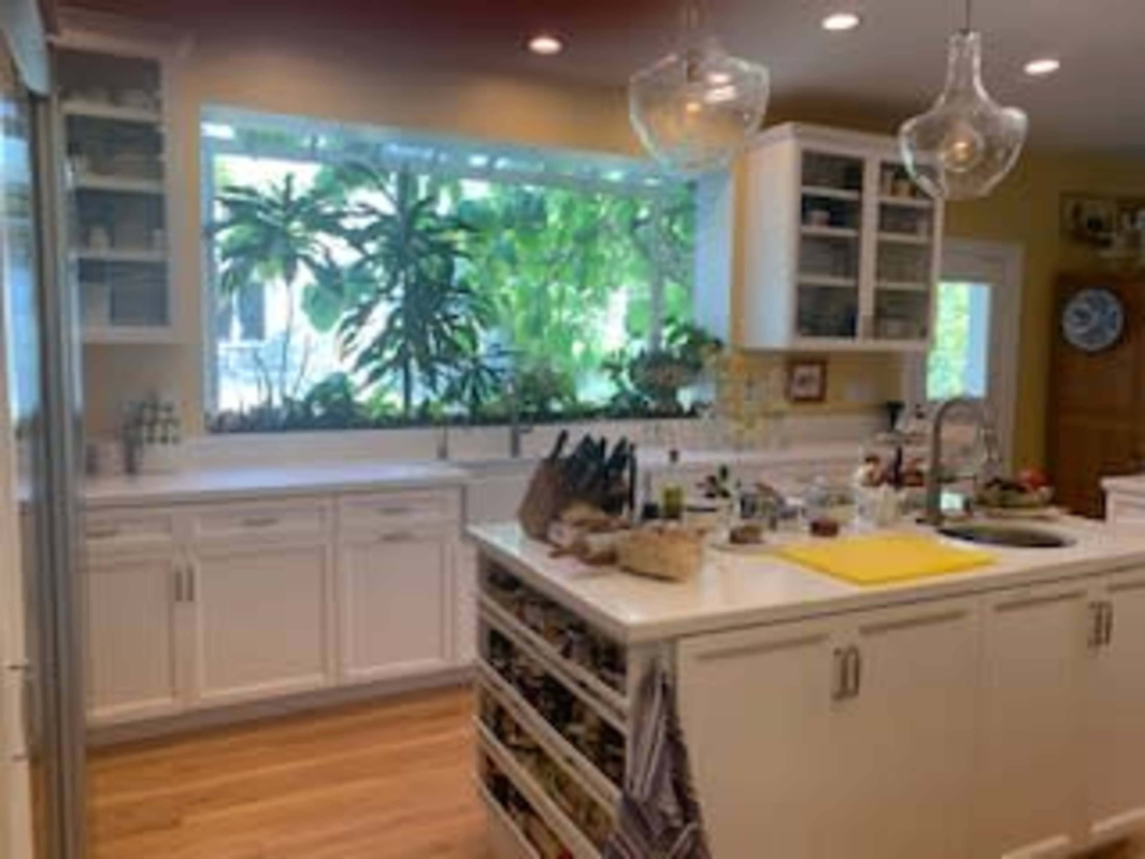 A bright kitchen features white cabinetry, a central island with storage, and large windows displaying lush green plants.