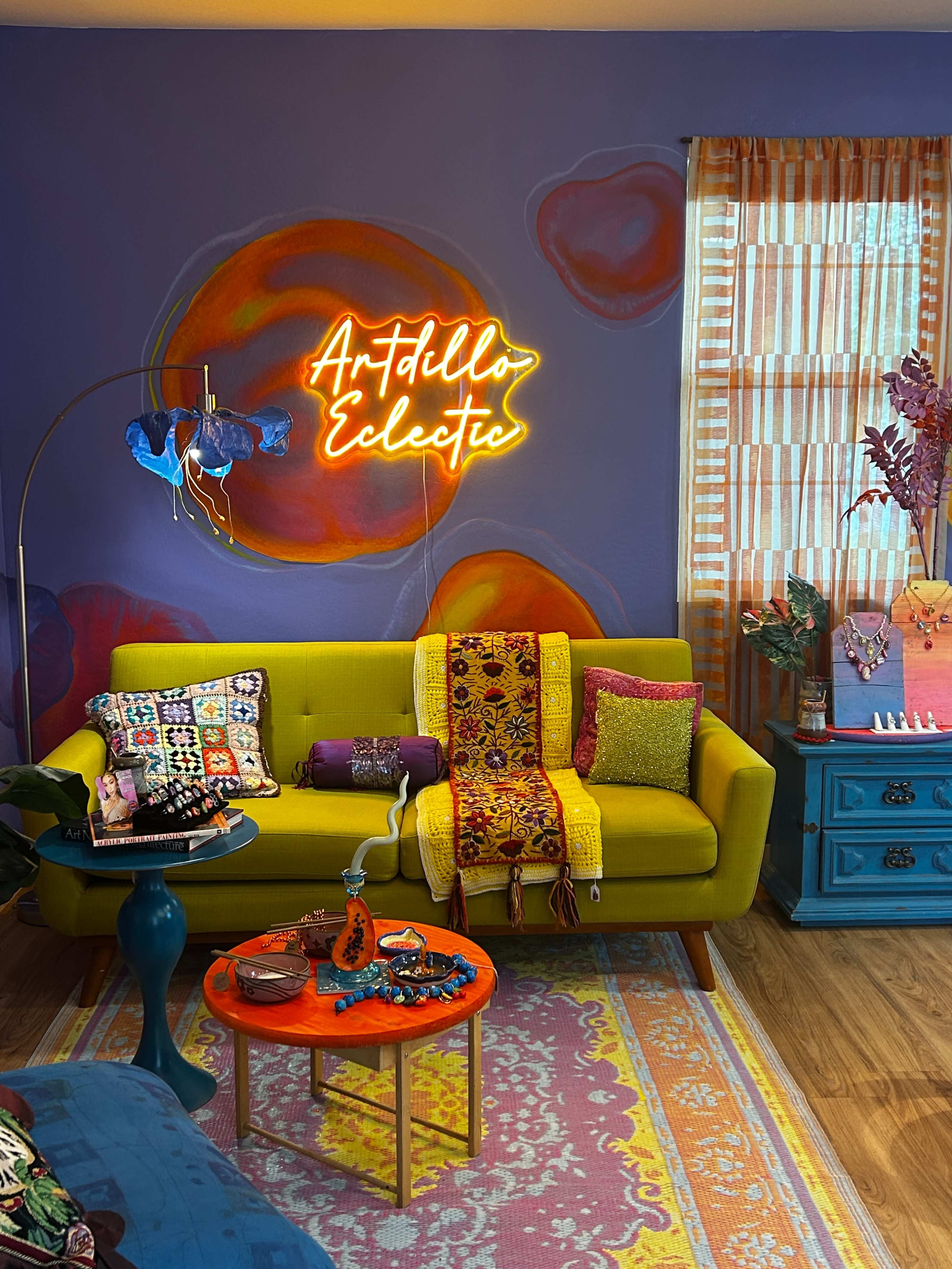 The image shows a vibrant living room featuring a yellow sofa adorned with colorful cushions, a neon sign that says "Artful Eclectic," and a small coffee table with various decorative items.