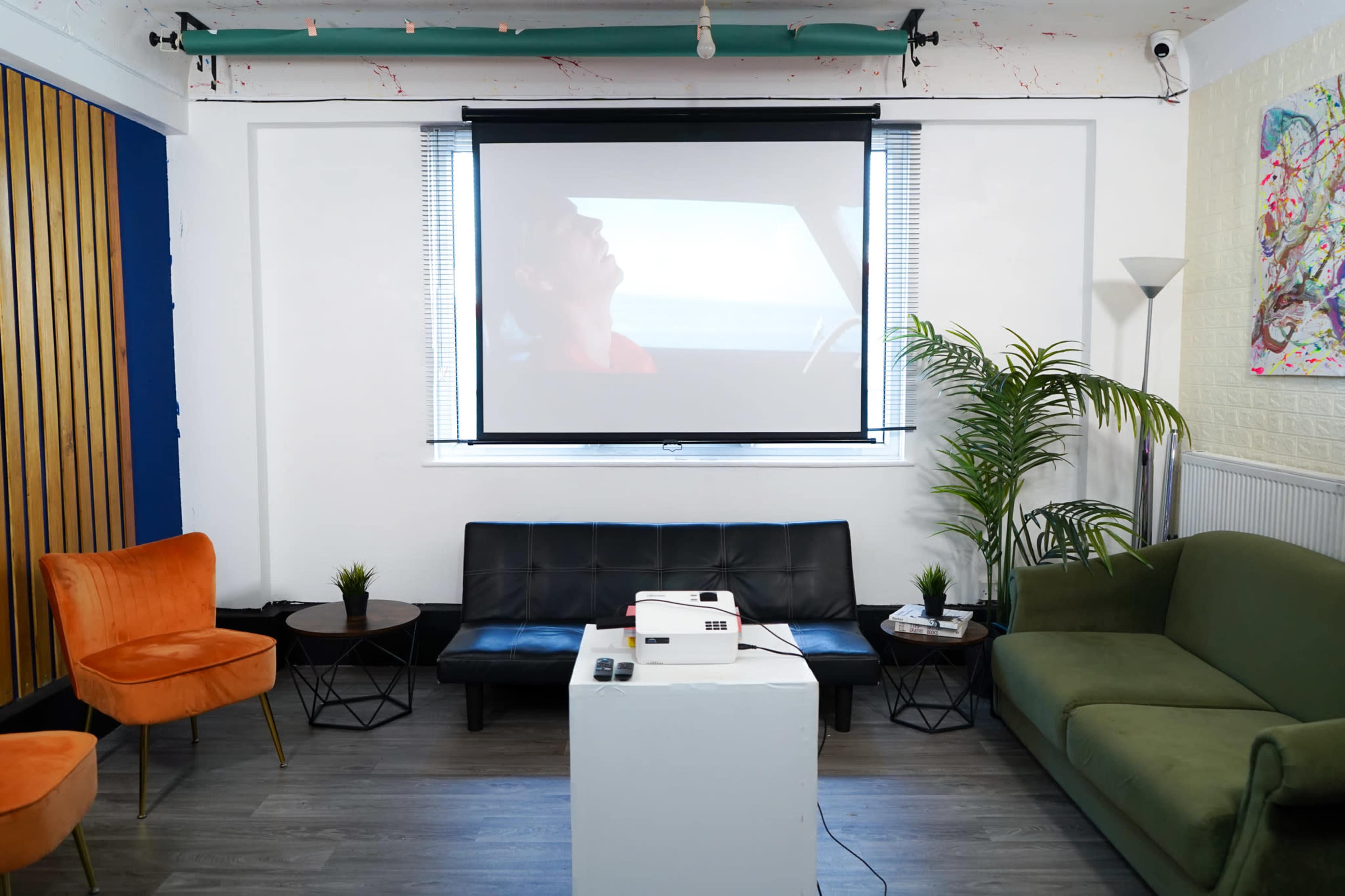A modern living room setup with a projector displaying a film on a screen, surrounded by various seating arrangements and decorative plants.