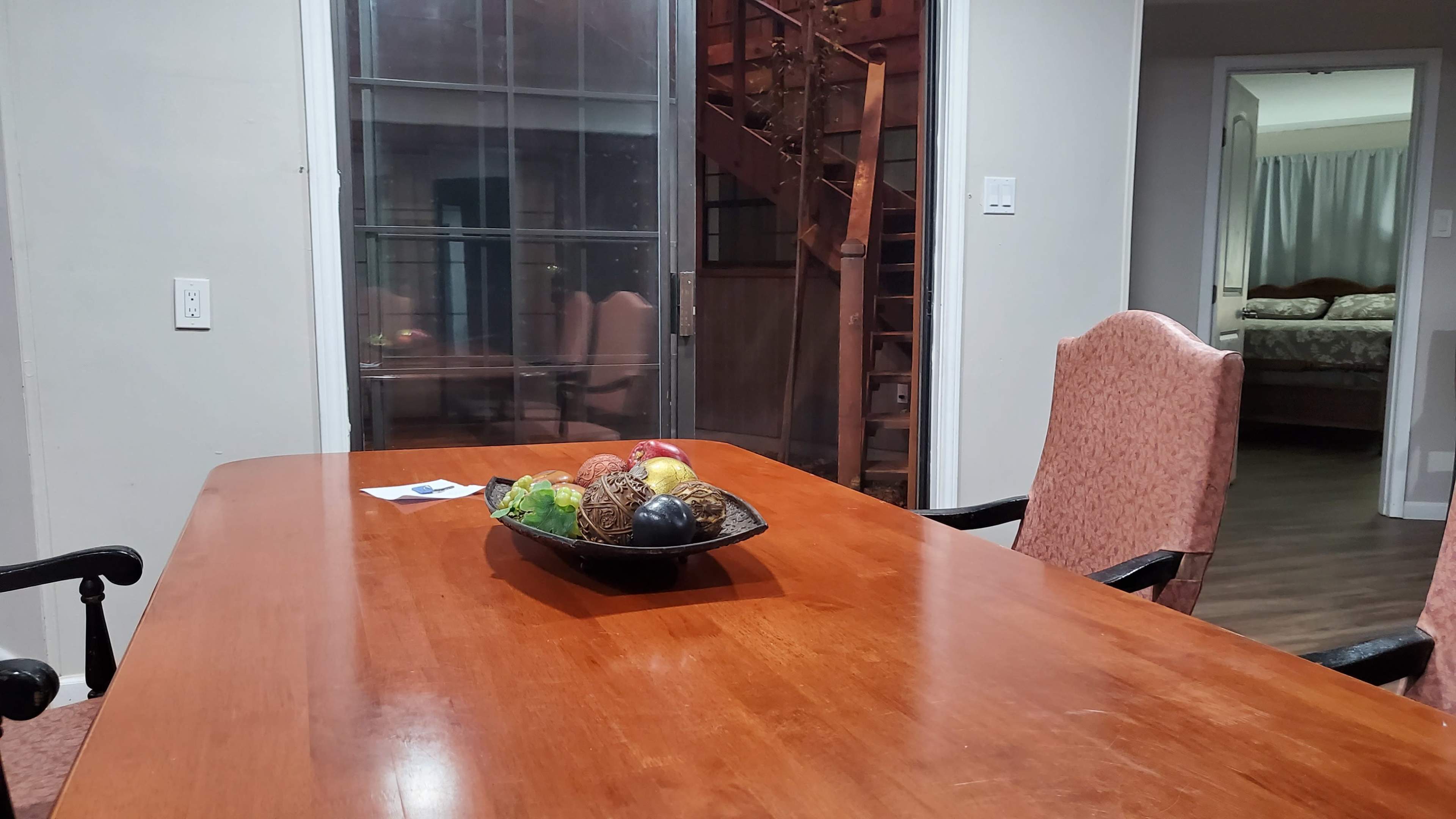A wooden dining table with a decorative bowl of fruit sits in a room with glass doors leading to a staircase and another room.